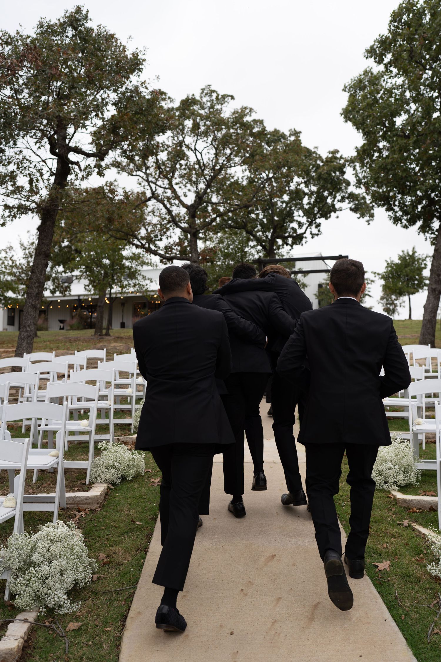 Wedding party in black suits walking down a path lined with white chairs.