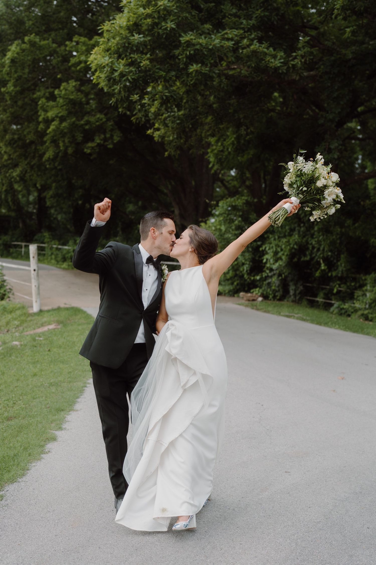 Newlyweds share an enthusiastic celebration moment on their wedding day along a tree-lined path.