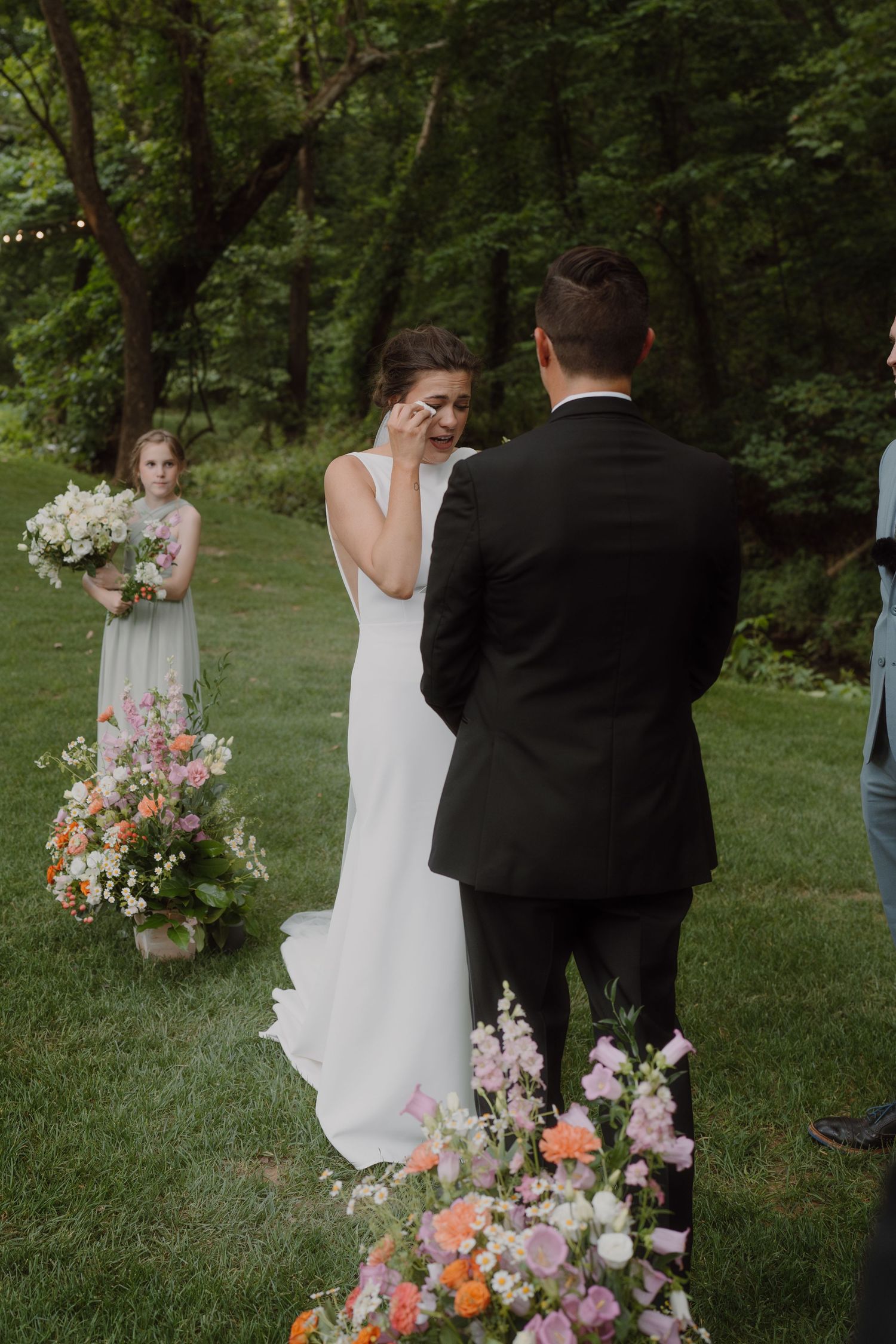 Outdoor wedding ceremony with floral arrangements and white dress against lush green background.
