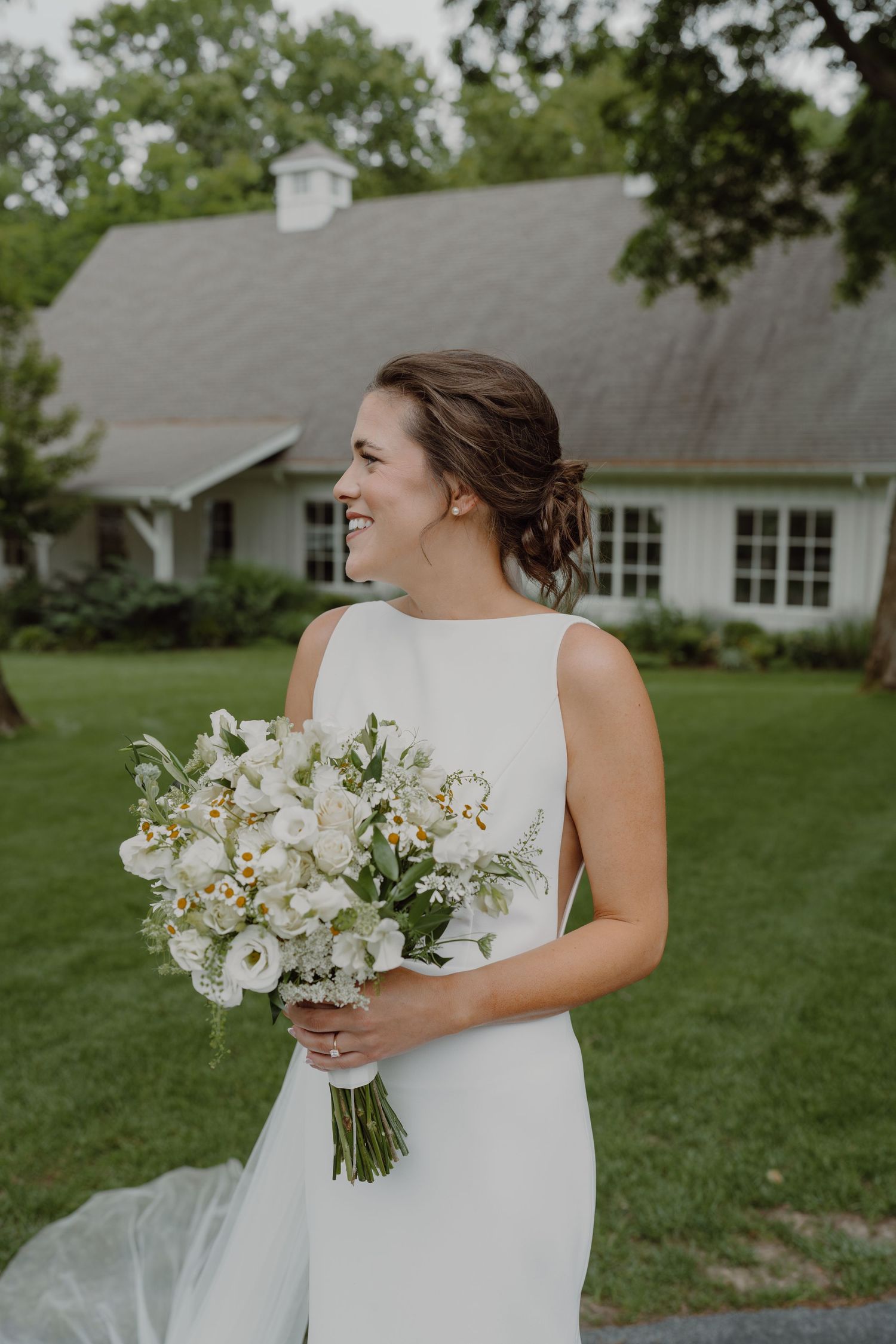 A bride in a classic white dress holds a bouquet of white flowers on a lawn near a building.