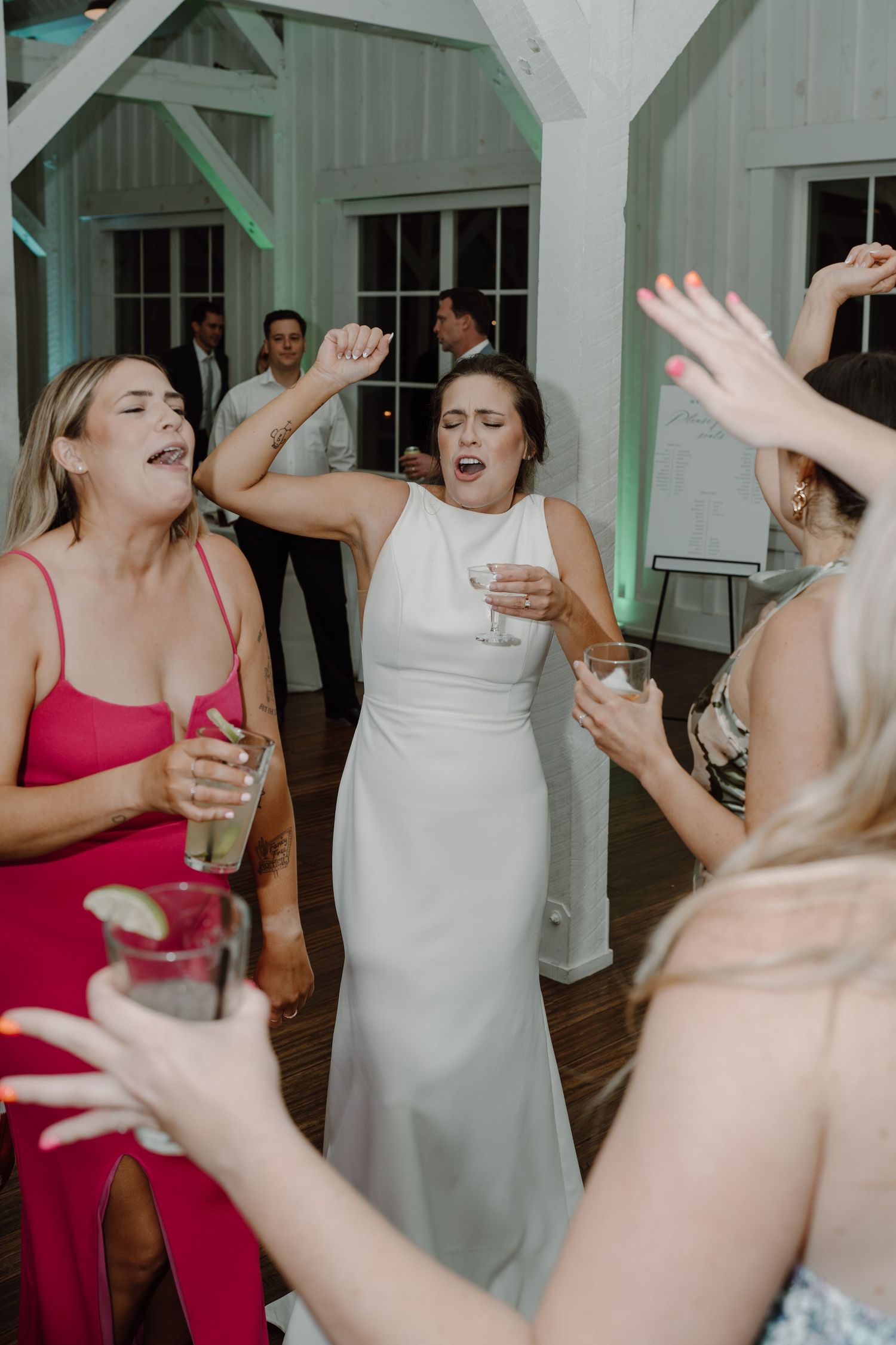 Guests in formal attire dance enthusiastically at a wedding reception with white interior.