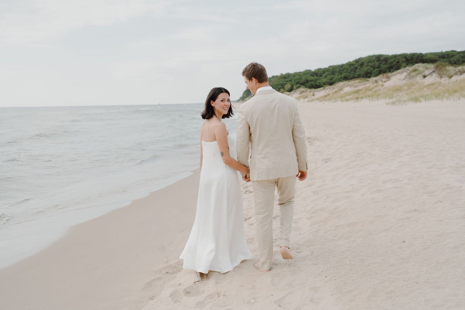 A couple in white and beige clothing walks together on a scenic coastal beach.