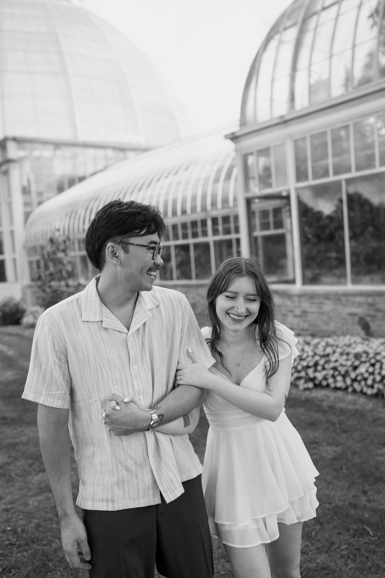 A romantic couple shares playful moments in front of a greenhouse with pink flowers blooming in the background.