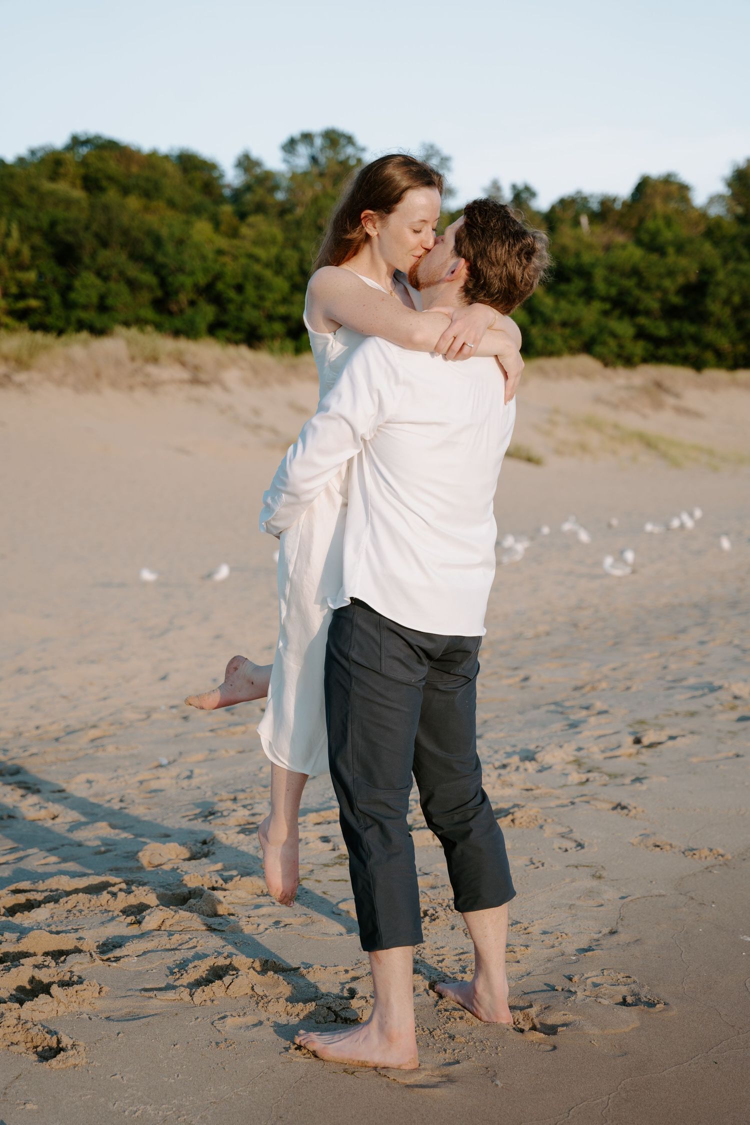Couple in casual attire sharing tender moments on beach with seagulls nearby.