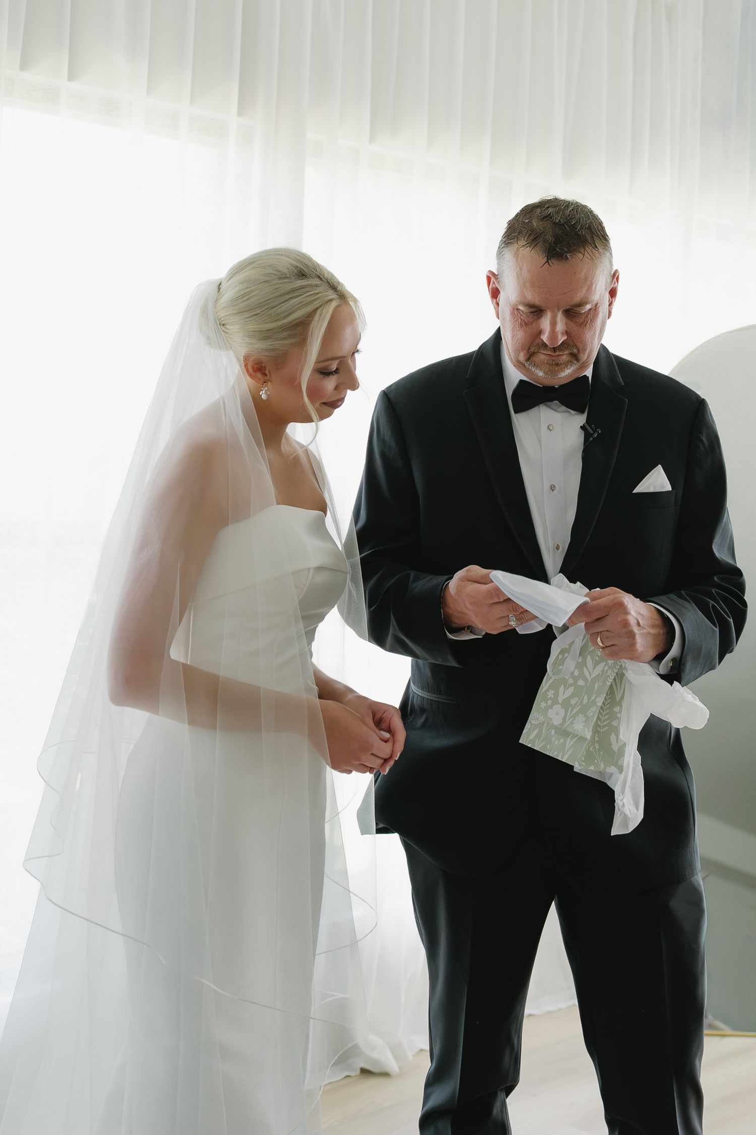 Bride in white gown and groom in black tuxedo reading wedding vows together indoors.