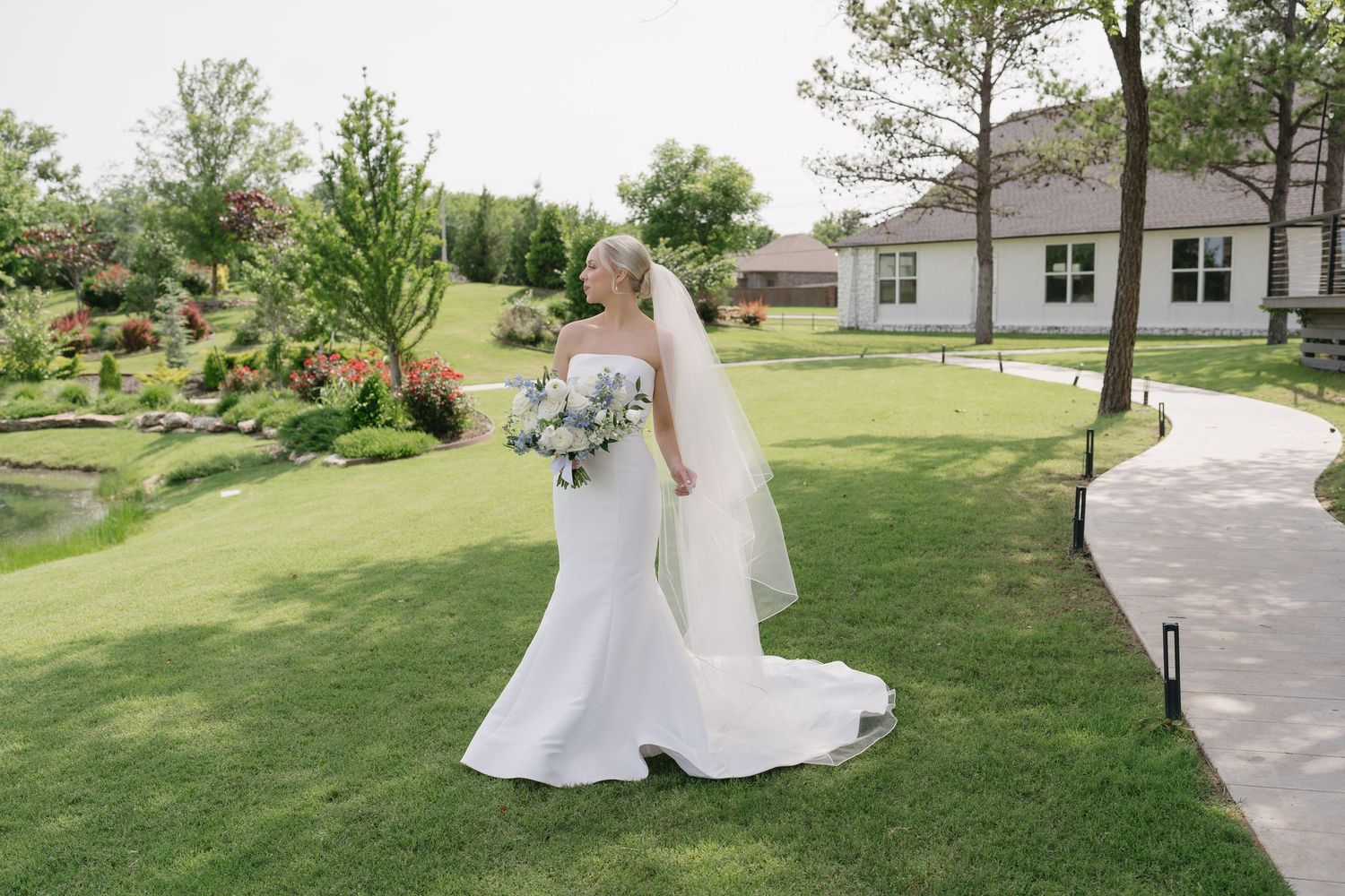 Bride in white strapless gown and veil holds blue bouquet on lush green lawn beside elegant venue.