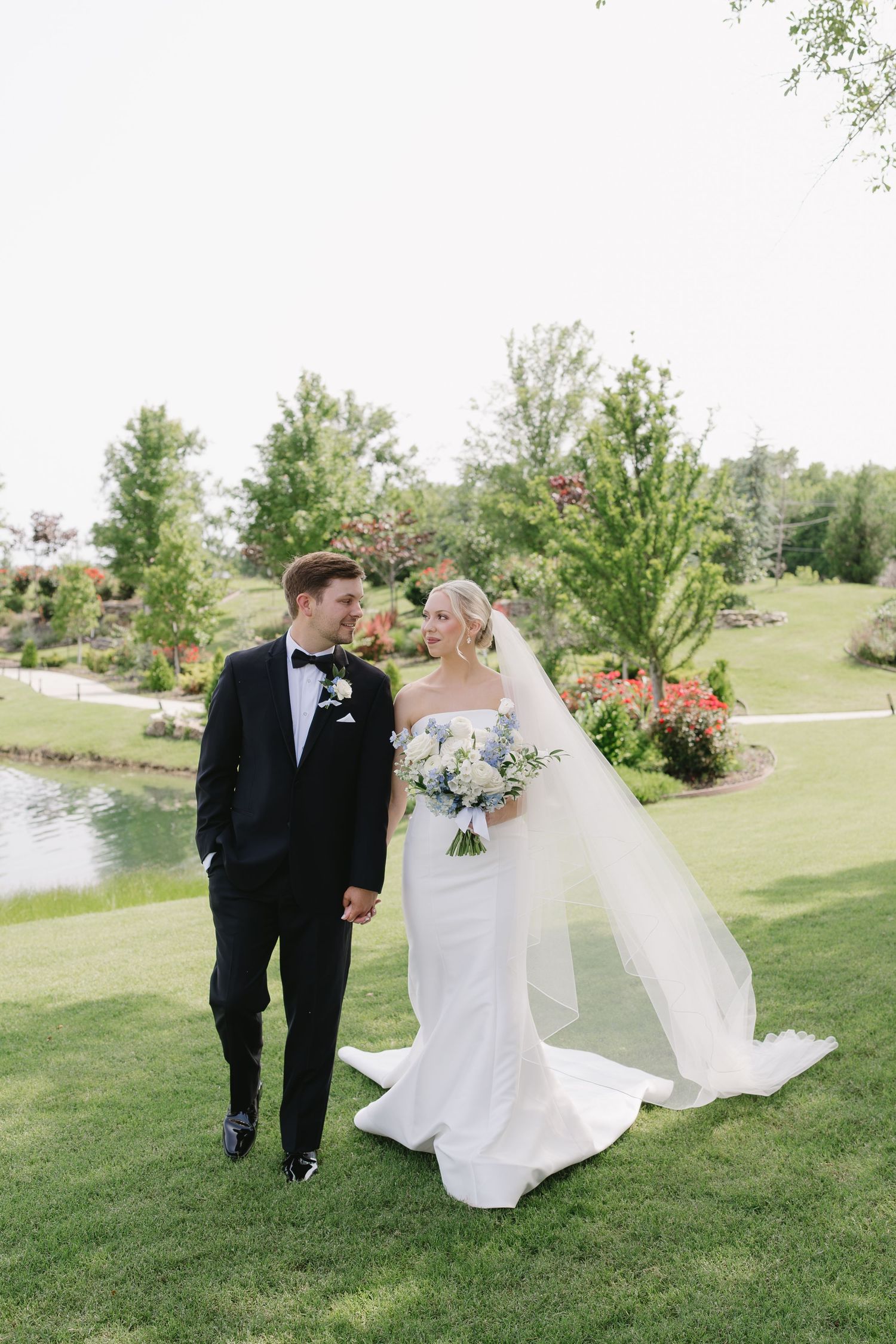 A bride and groom walk together on lush green grounds, she in a white gown with veil, he in a black tuxedo.