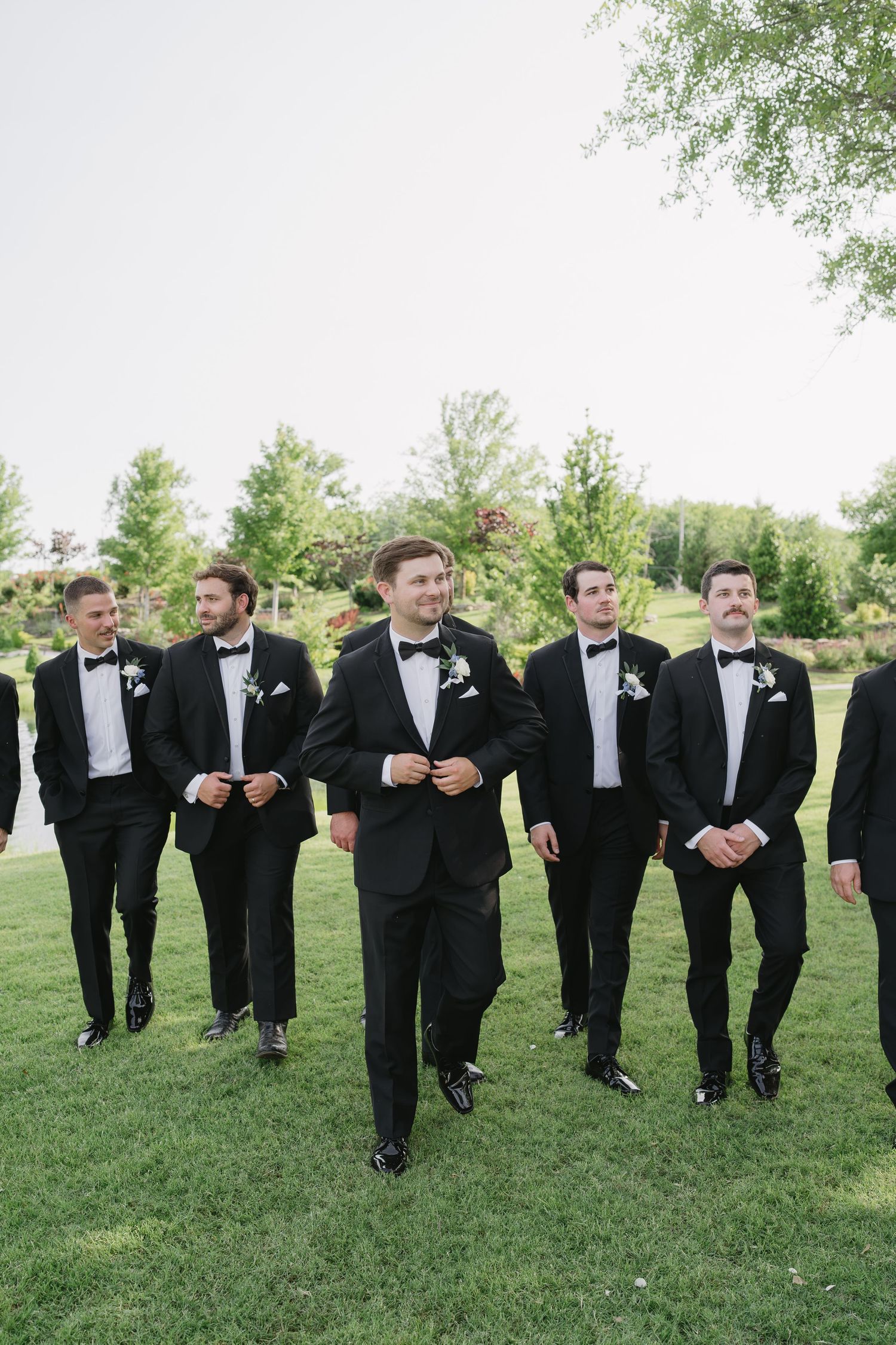 Groom and groomsmen in black tuxedos with bow ties and boutonnieres walking outdoors on green grass at a wedding.