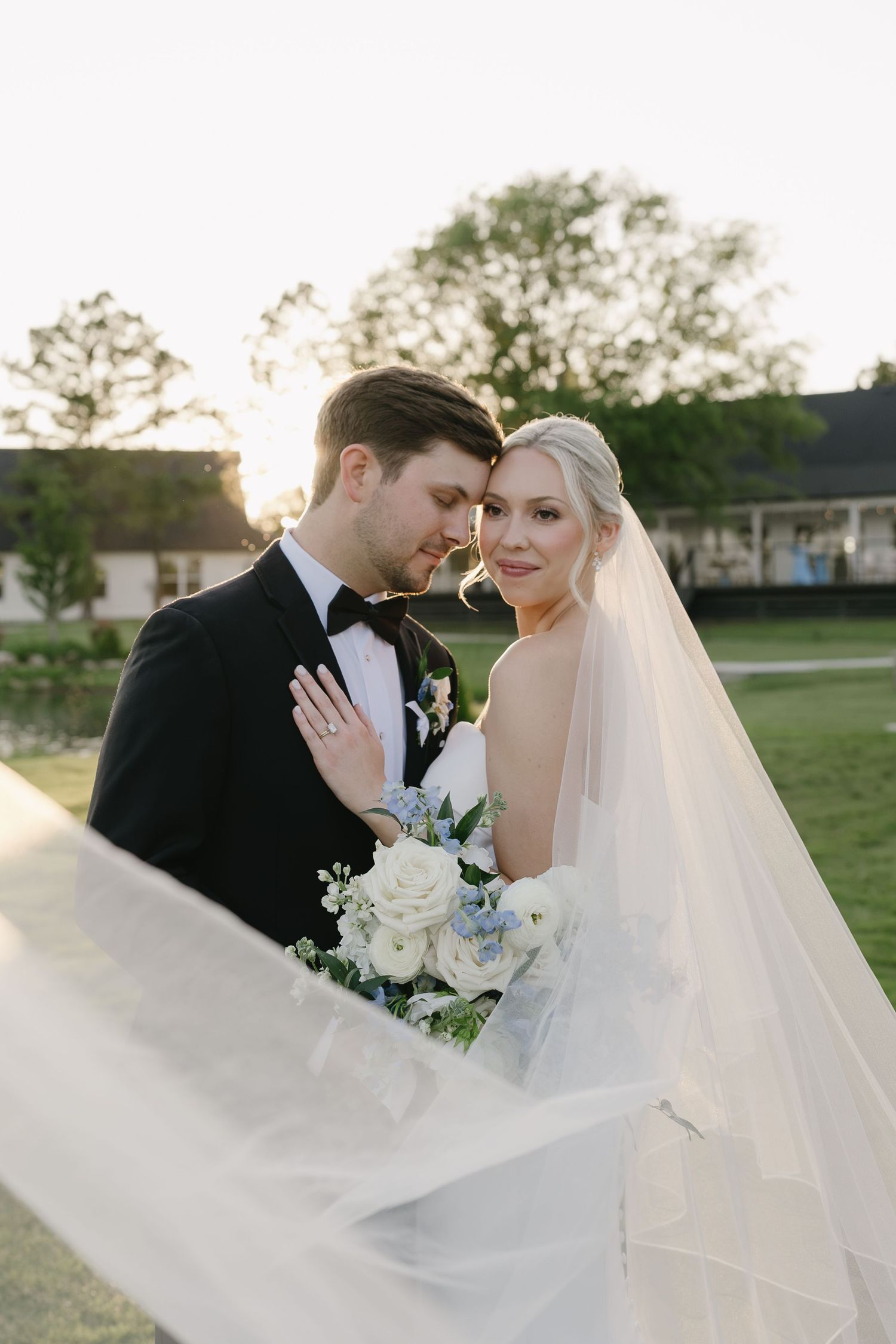 Bride and groom share an intimate moment at golden hour, her veil flowing as she holds a white and blue bouquet.