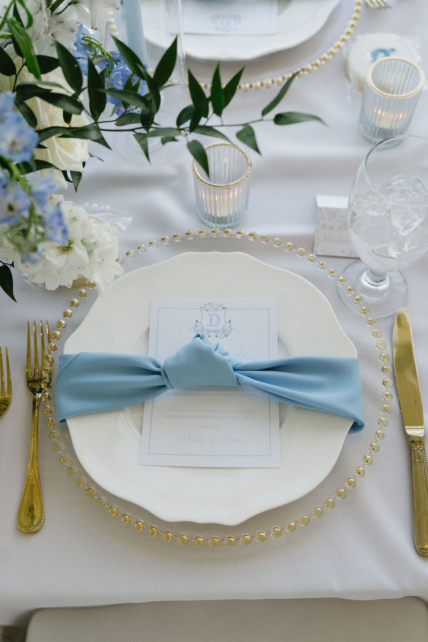 Elegant wedding place setting with blue bow napkin, gold flatware, pearl charger plate, and blue floral centerpiece on white linen.