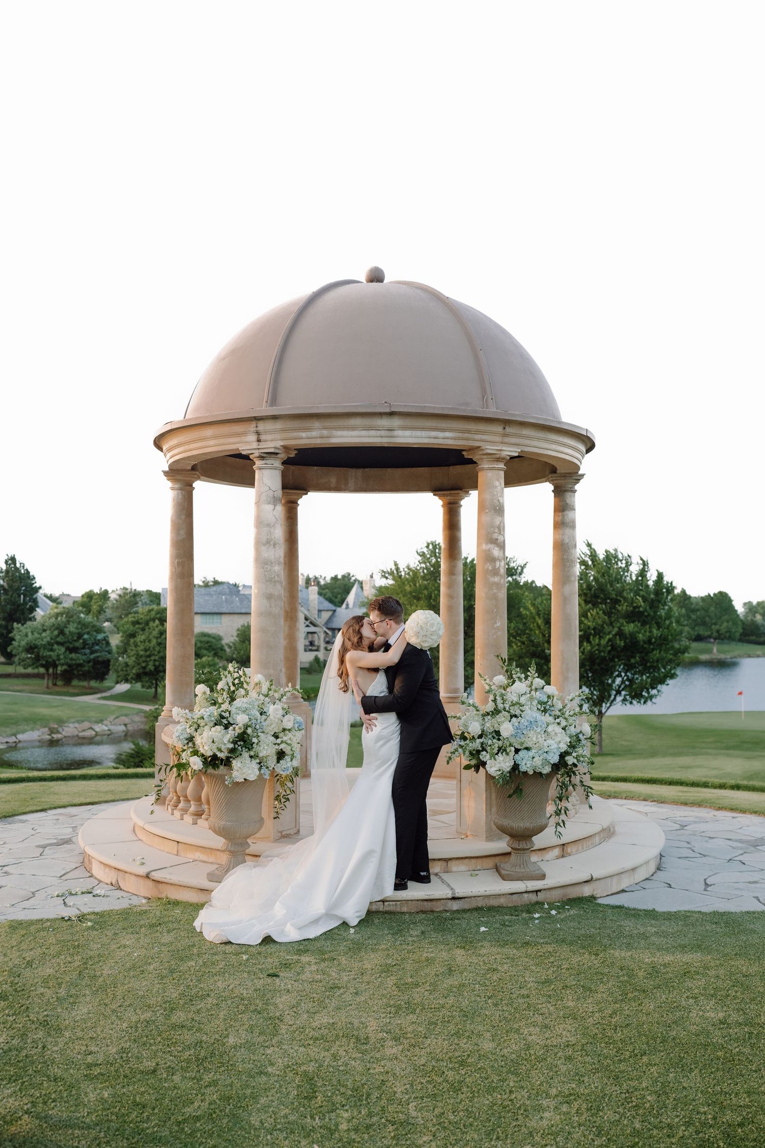 A romantic wedding couple shares intimate moments in a beautiful garden gazebo surrounded by white floral arrangements.
