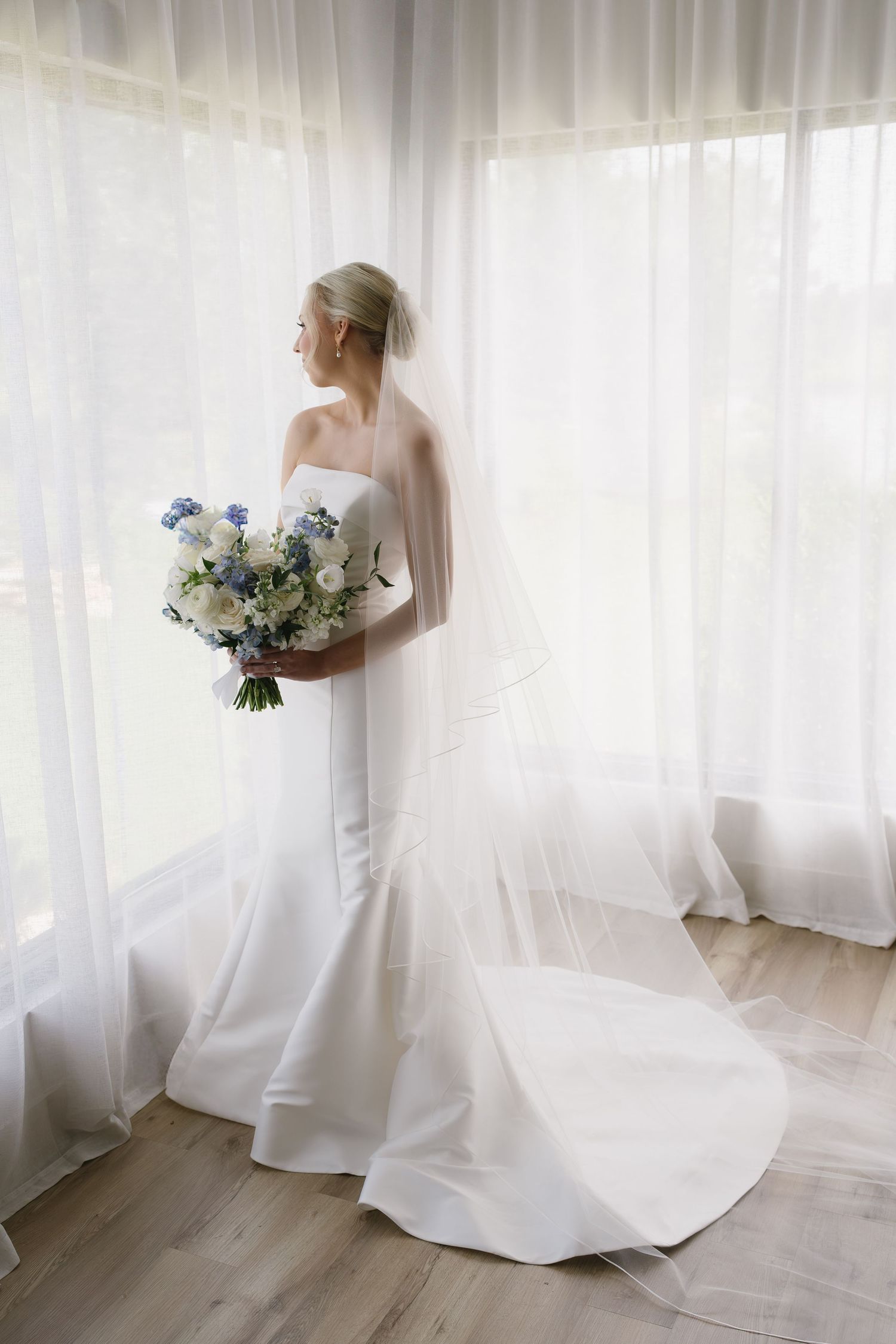 Elegant bride in white strapless gown with veil holding blue and white bouquet, standing by sheer curtained windows.