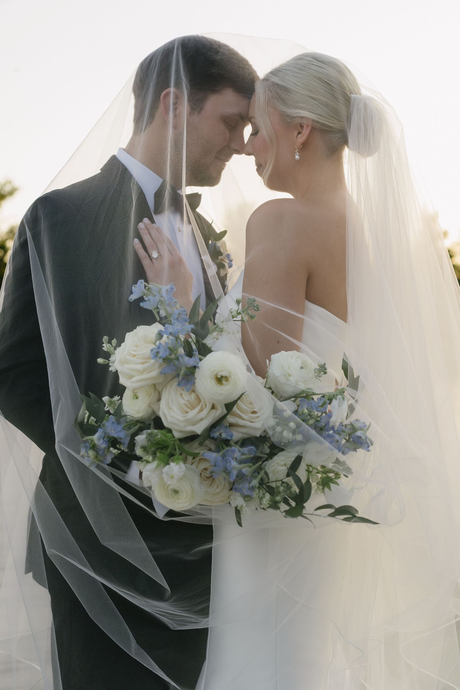 Bride and groom share an intimate moment under a flowing veil, holding a white and blue floral bouquet.