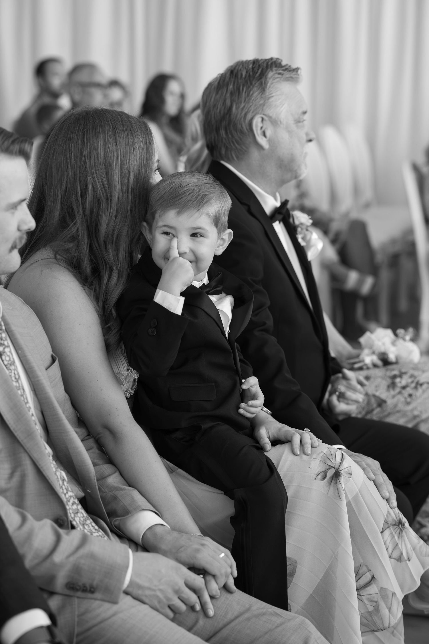 Black and white photo of a young boy in a tuxedo sitting with wedding guests during a ceremony.
