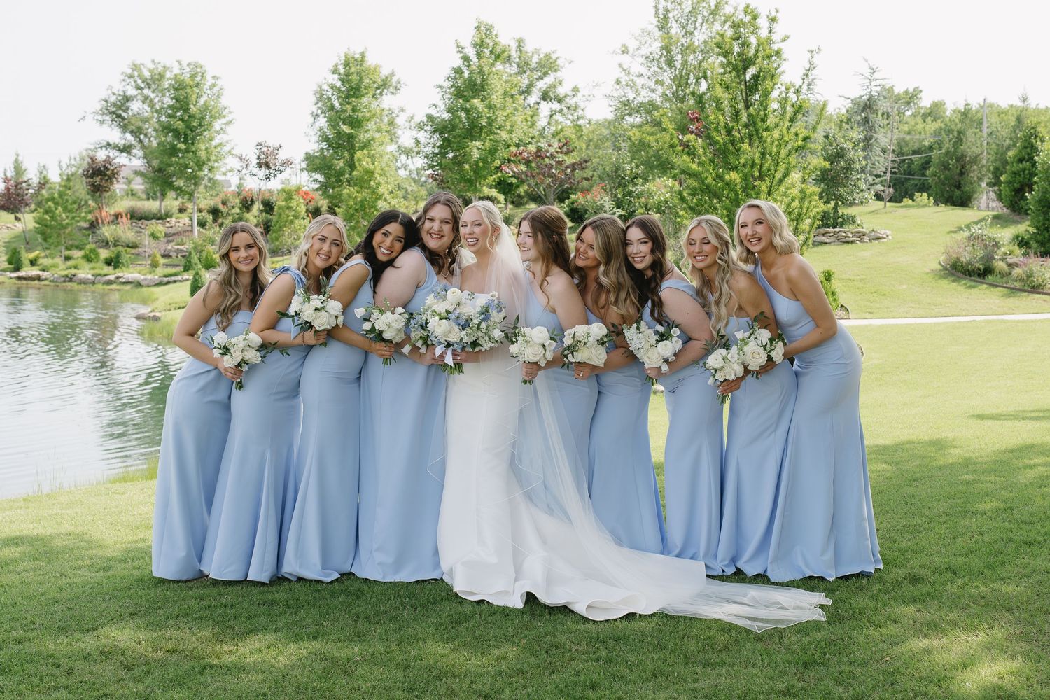Bride and bridesmaids in light blue gowns holding white bouquets, smiling outdoors near a pond on a sunny day.