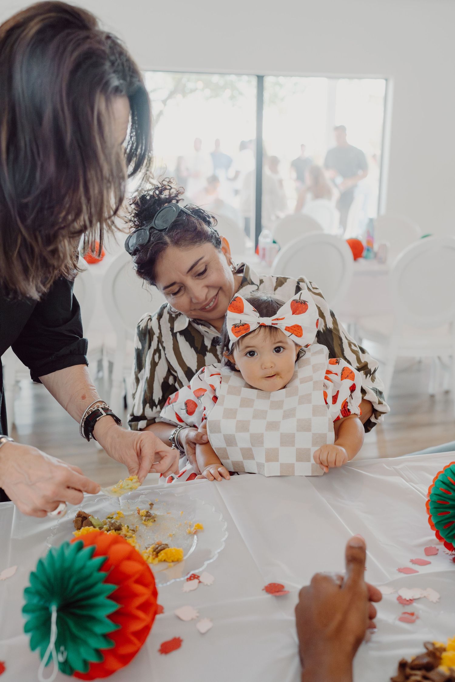 Baby sitting at white table with colorful party decorations and orange tissue paper flowers.
