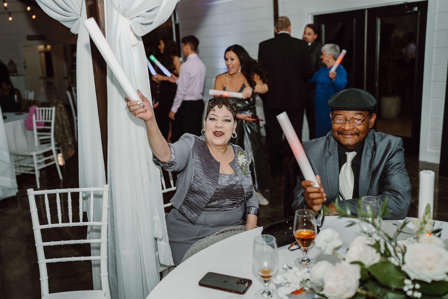 Wedding guests enjoy a lively moment at a reception table decorated with white linens and floral centerpieces.