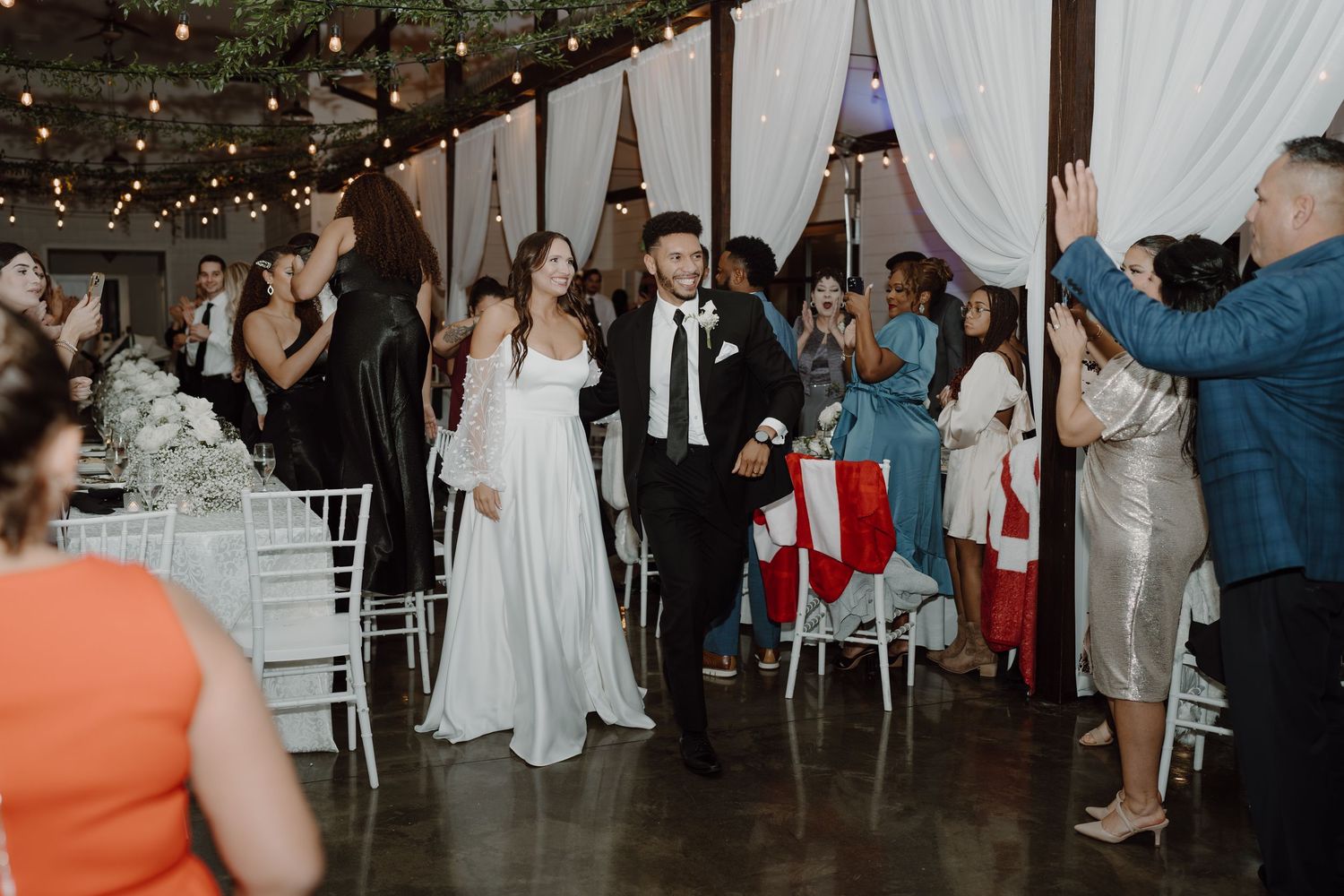 Newlyweds make their grand entrance at indoor reception while guests cheer and celebrate.