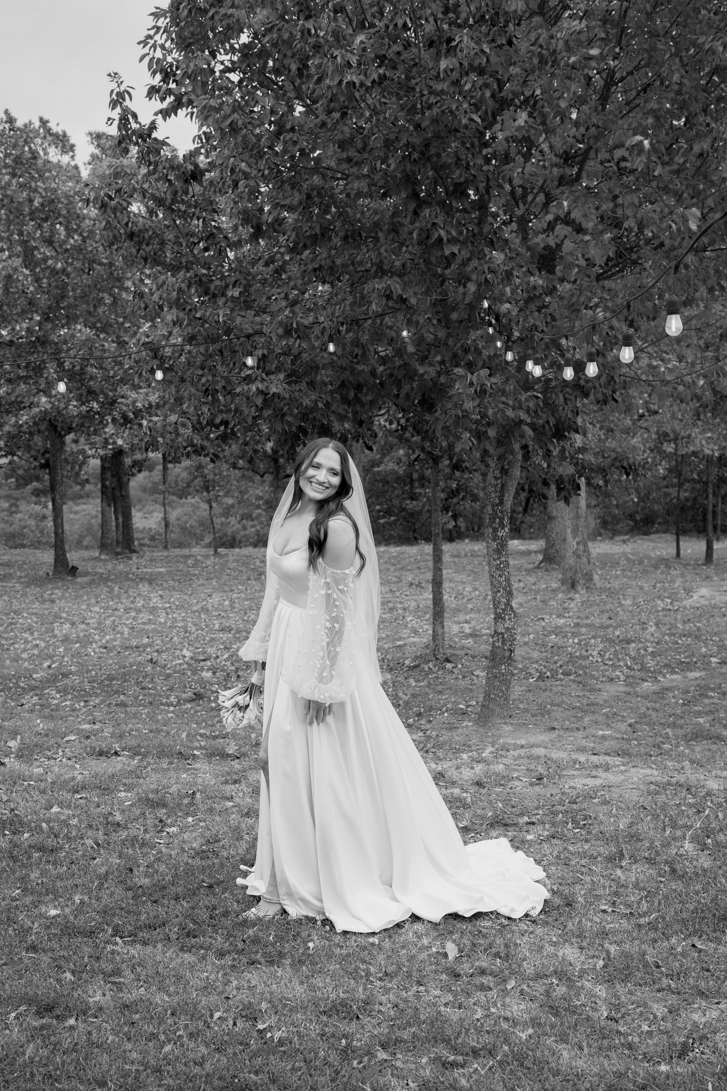 A black and white photo of a wedding dress with long sleeves in a garden setting with string lights.