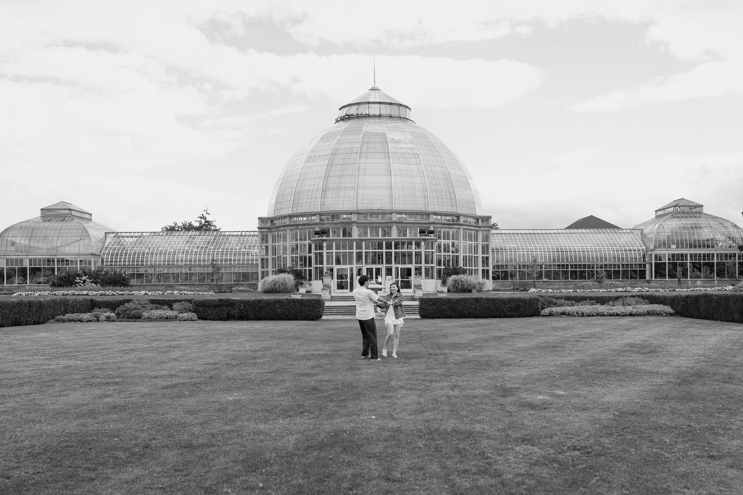 Black and white wide shot of couple standing on lawn in front of large domed conservatory building.