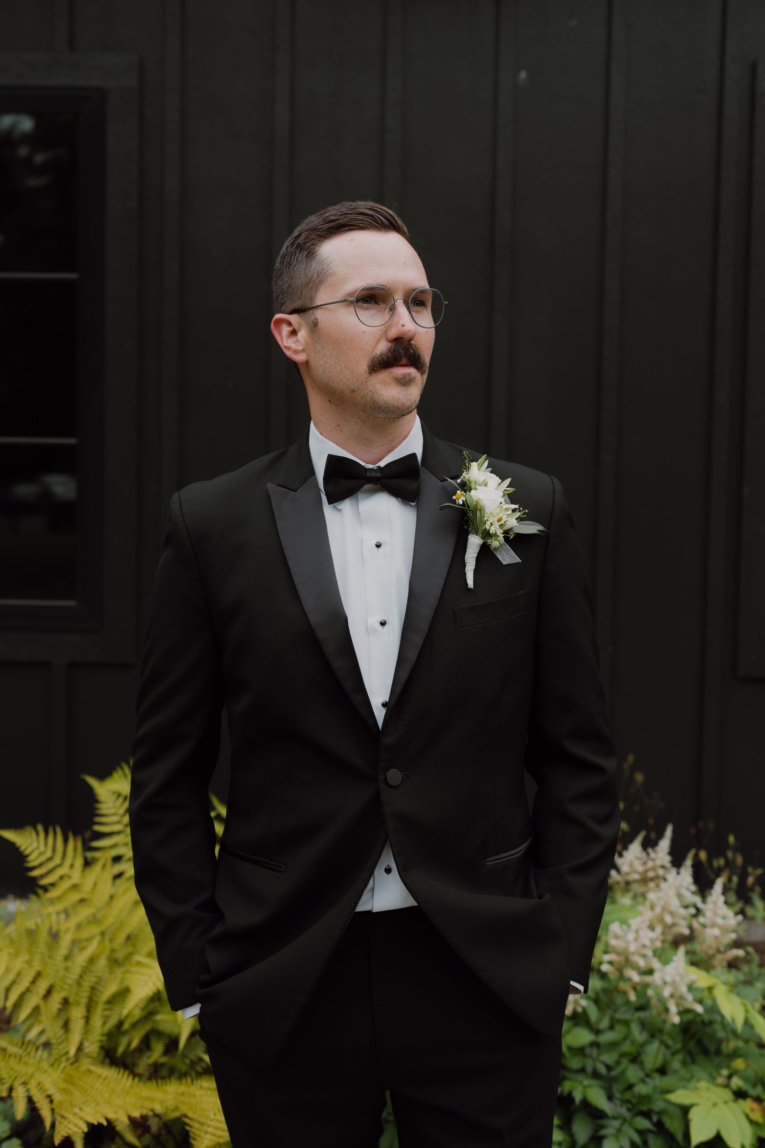 A person wearing glasses and a mustache poses in formal attire next to decorative plants.