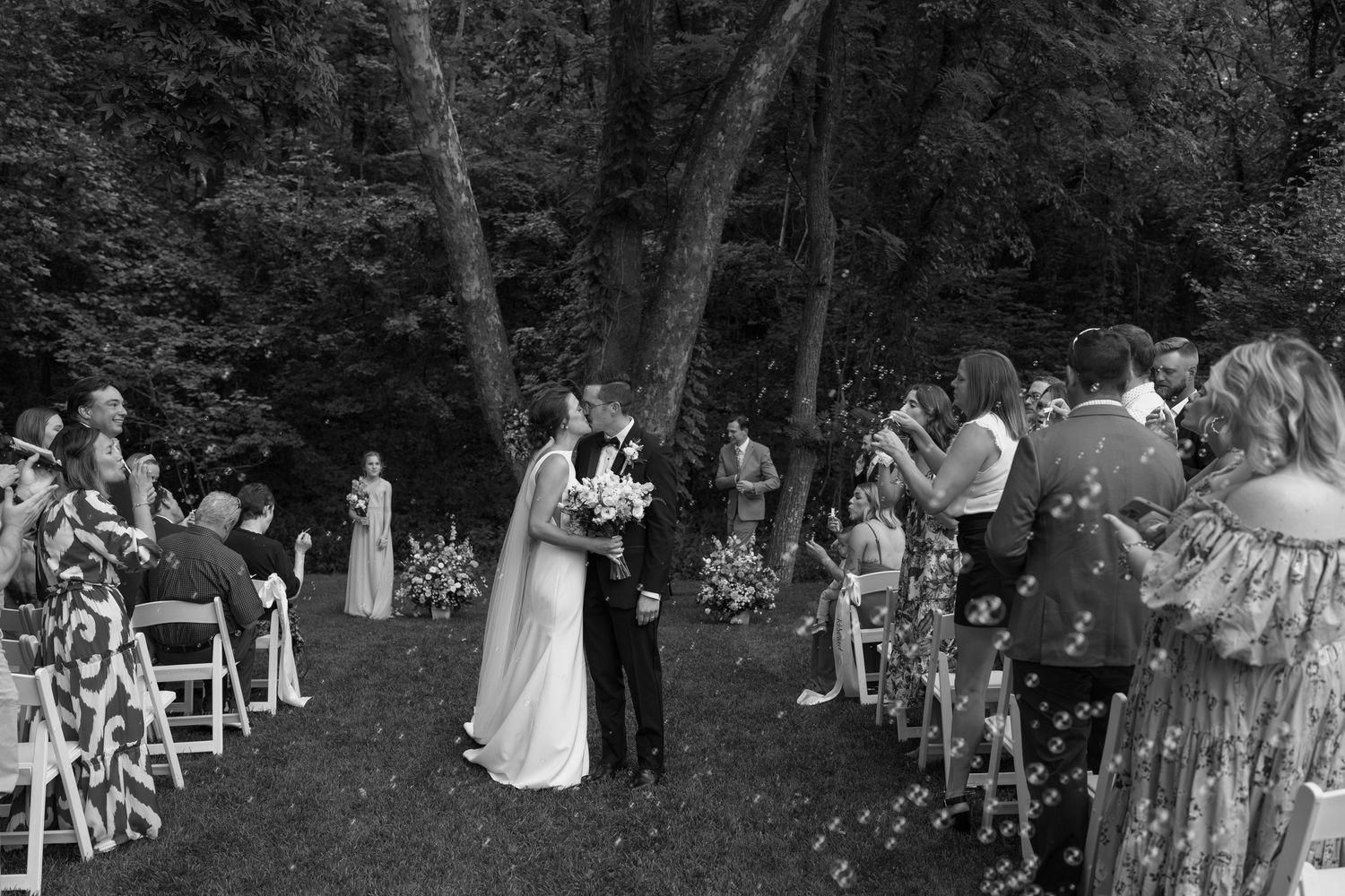Black and white photo of guests seated at an outdoor woodland wedding ceremony.