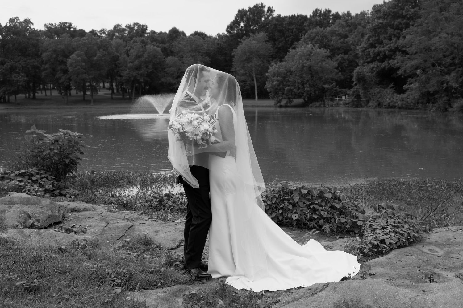 Black and white wedding photography by a serene lake with fountain.