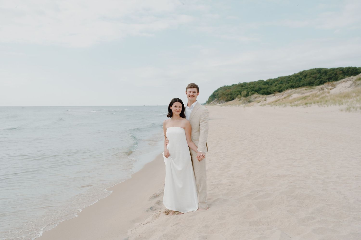 A couple stands together on a peaceful beach shoreline wearing formal attire.