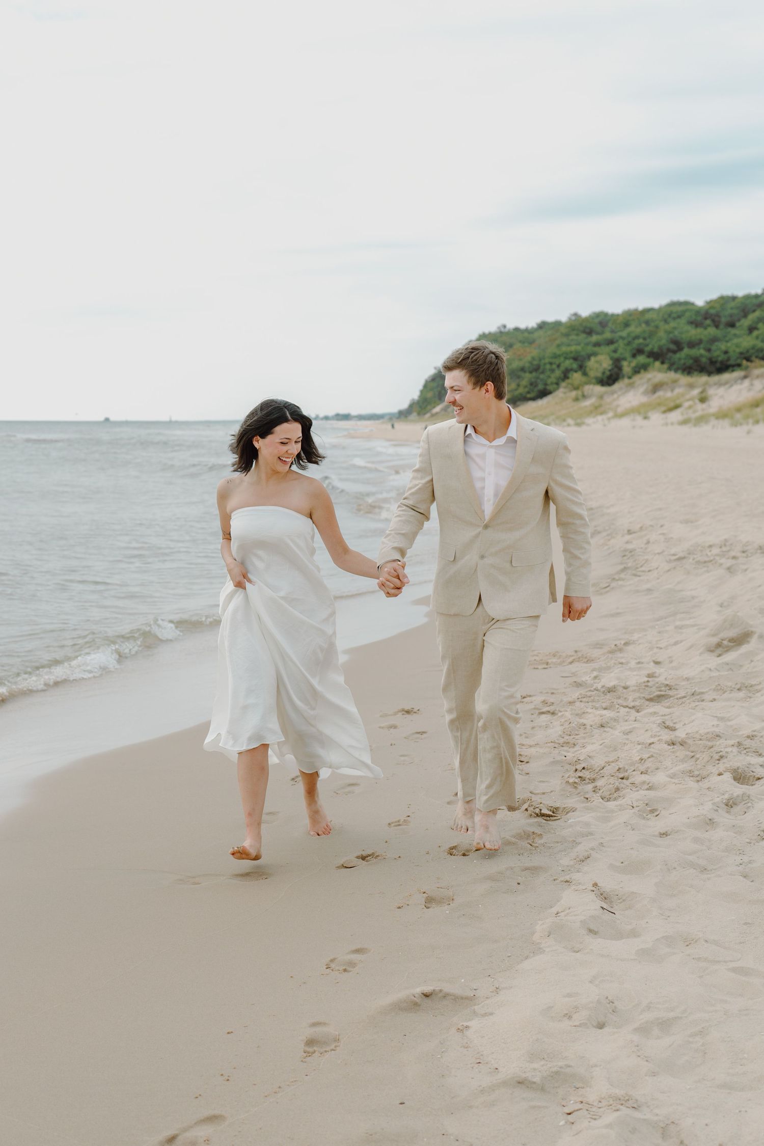 A couple in coordinated light outfits runs hand in hand along a scenic beachfront.