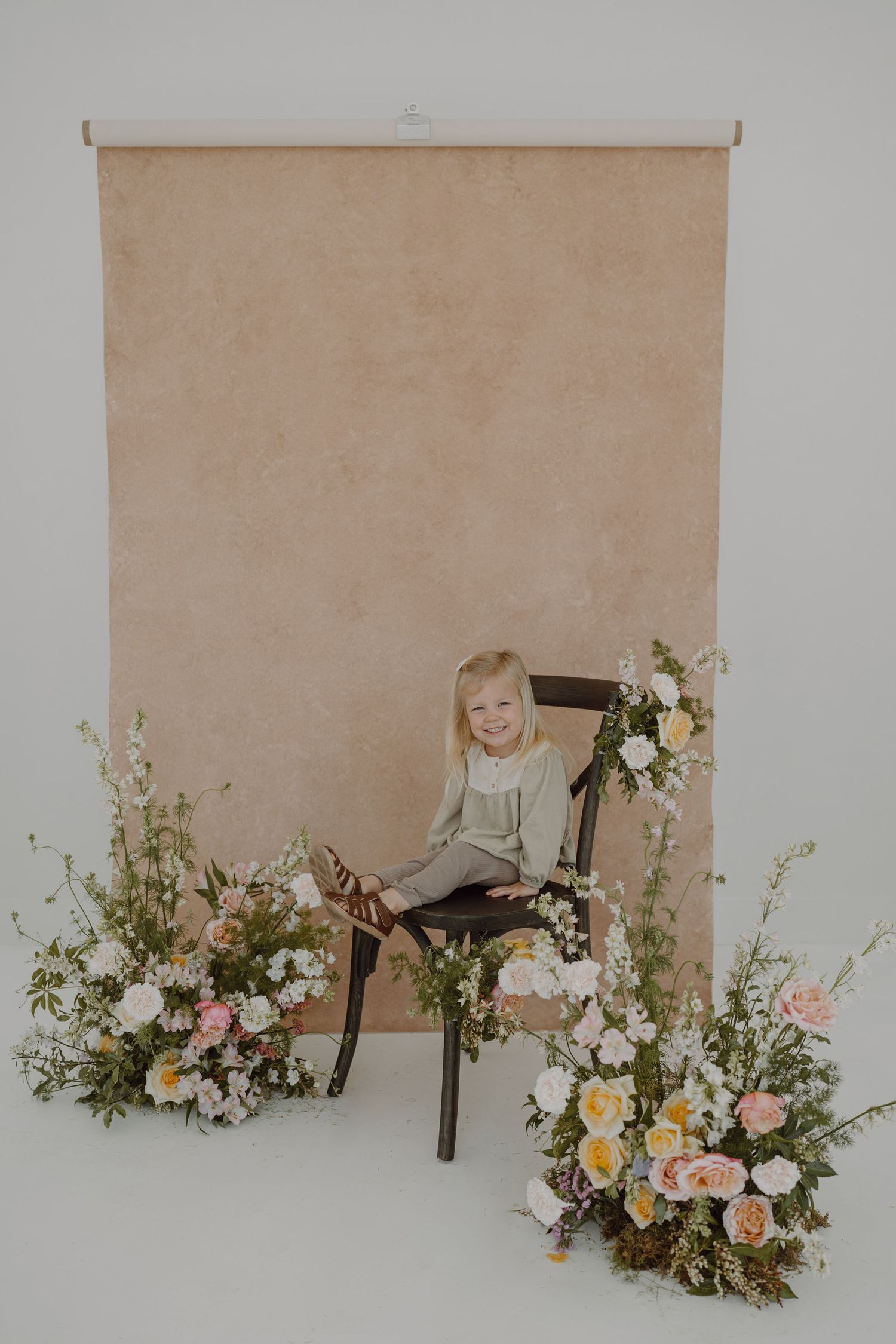 Portrait studio setup with elegant floral arrangements and wooden chair against neutral background.
