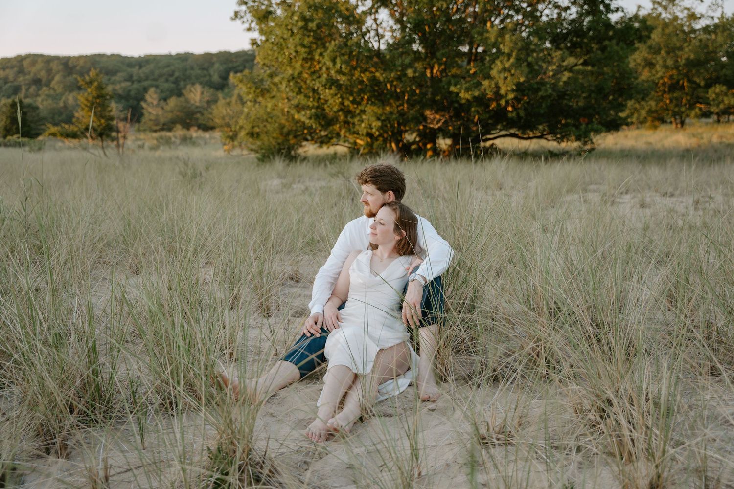 A romantic couple embraces in a golden field during sunset with trees in the background.