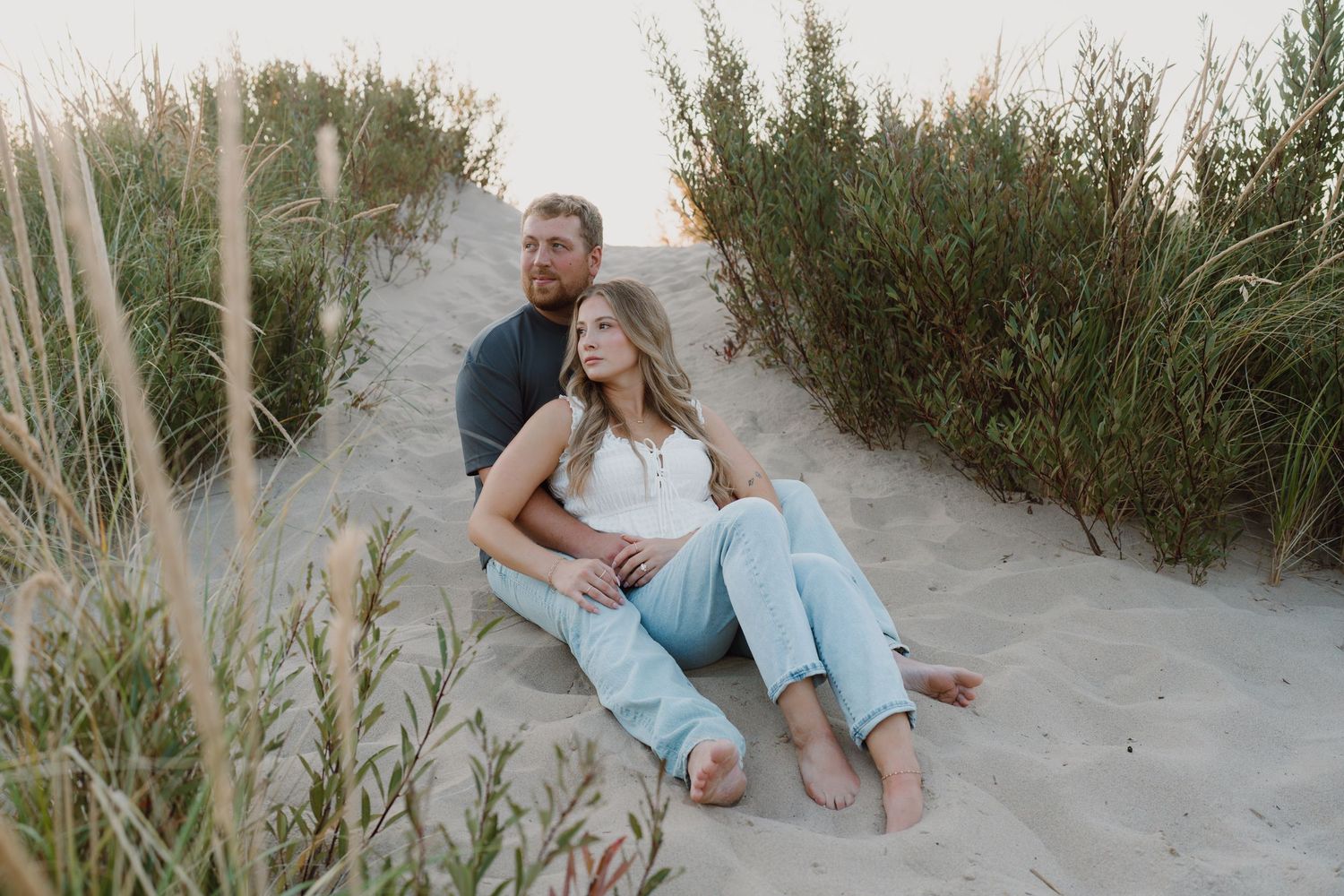 Two people share a romantic moment while sitting in the sand among beach grass.