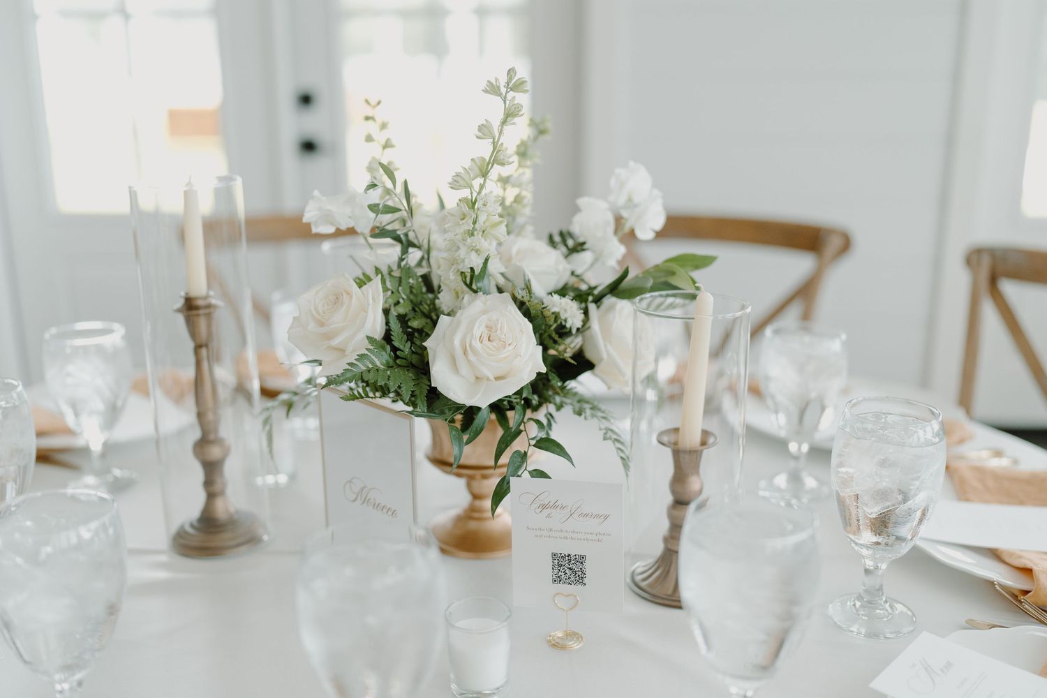 Minimalist white wedding reception table setting with delicate glassware and floral centerpieces.