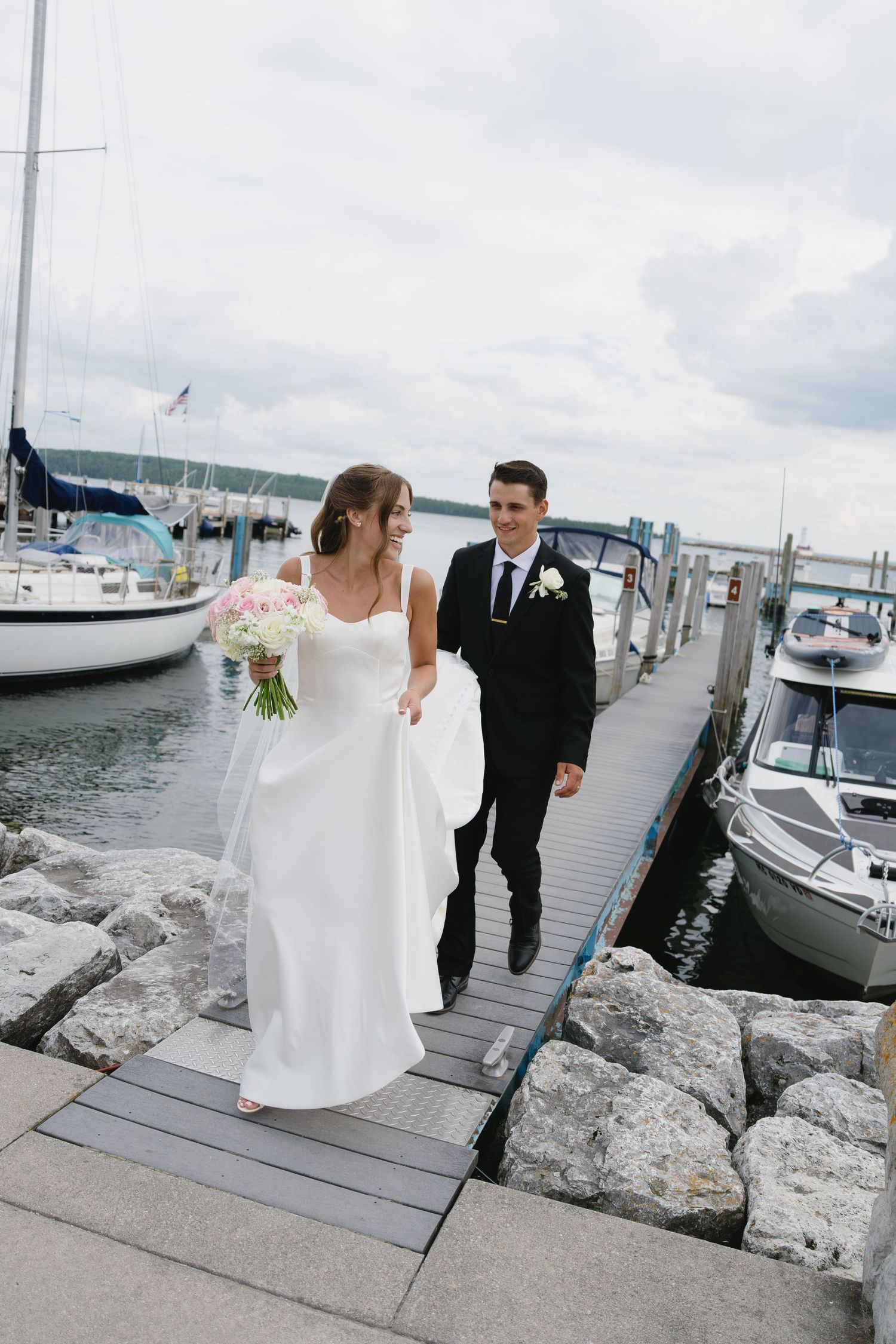 Bride and groom walk hand-in-hand on a marina dock, with sailboats and overcast skies in the background.