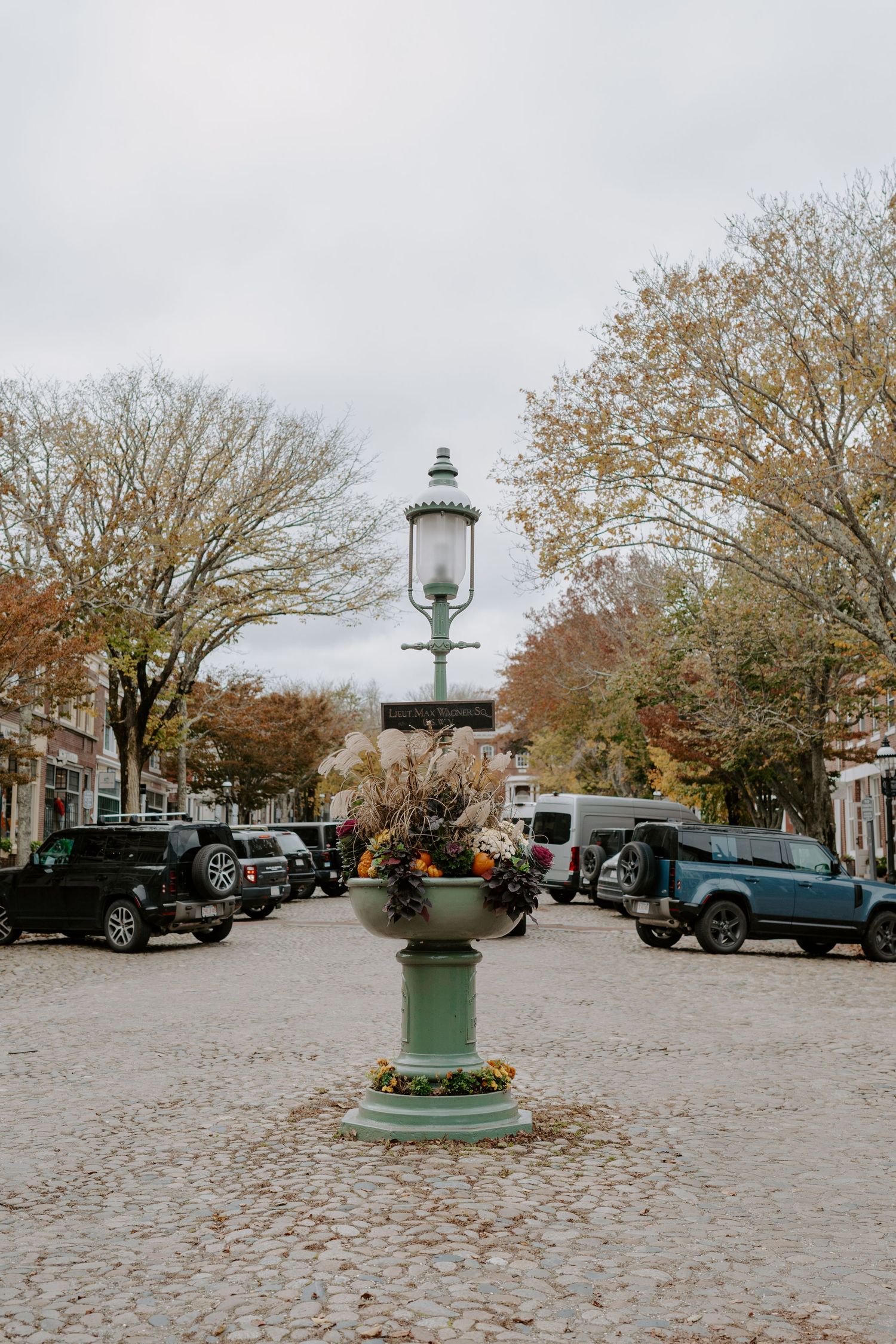 Historic cobblestone square with vintage lamp post and fall trees surrounded by classic cars.