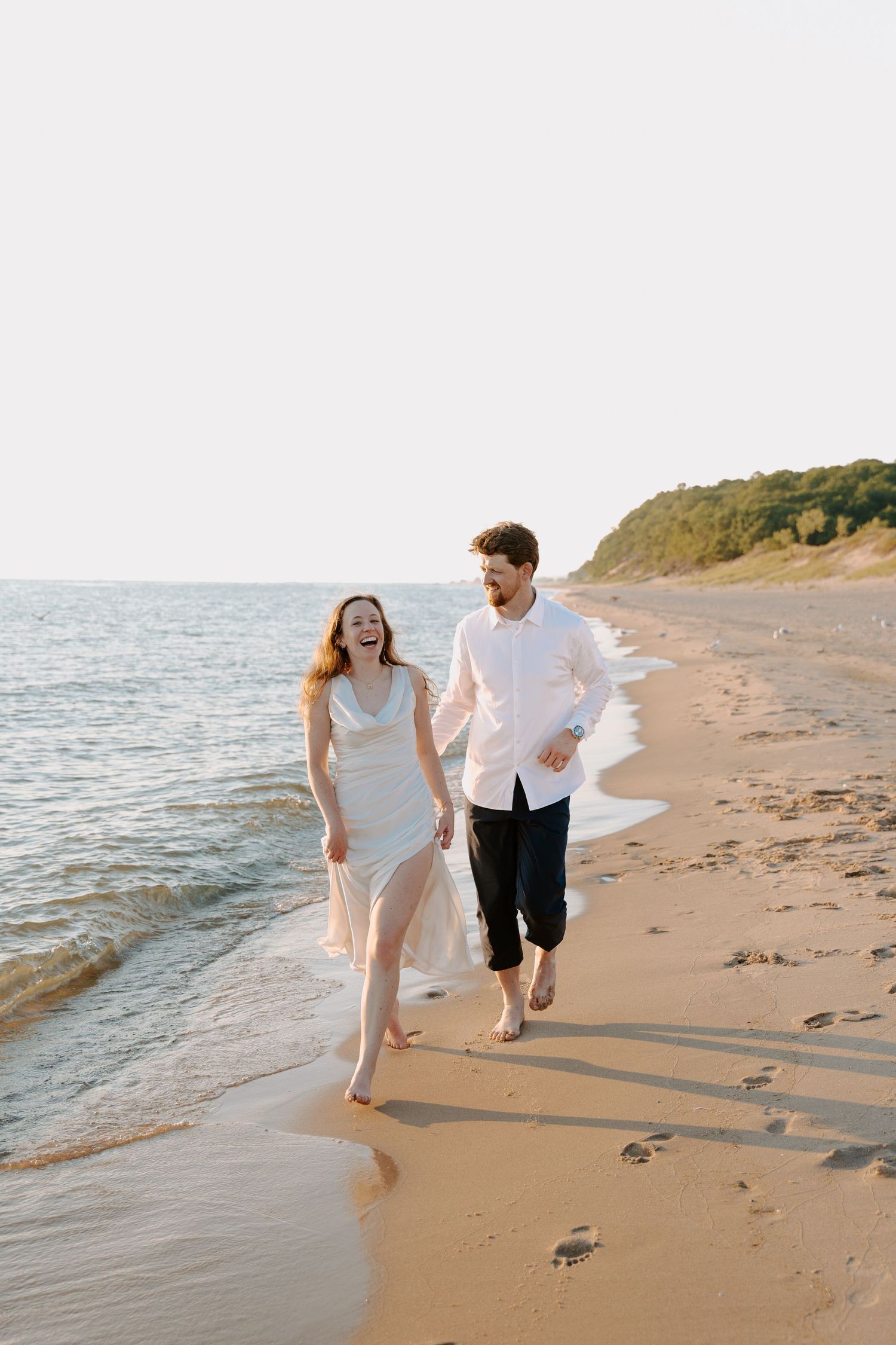 A couple runs playfully through the shallow waves on a sandy beach.