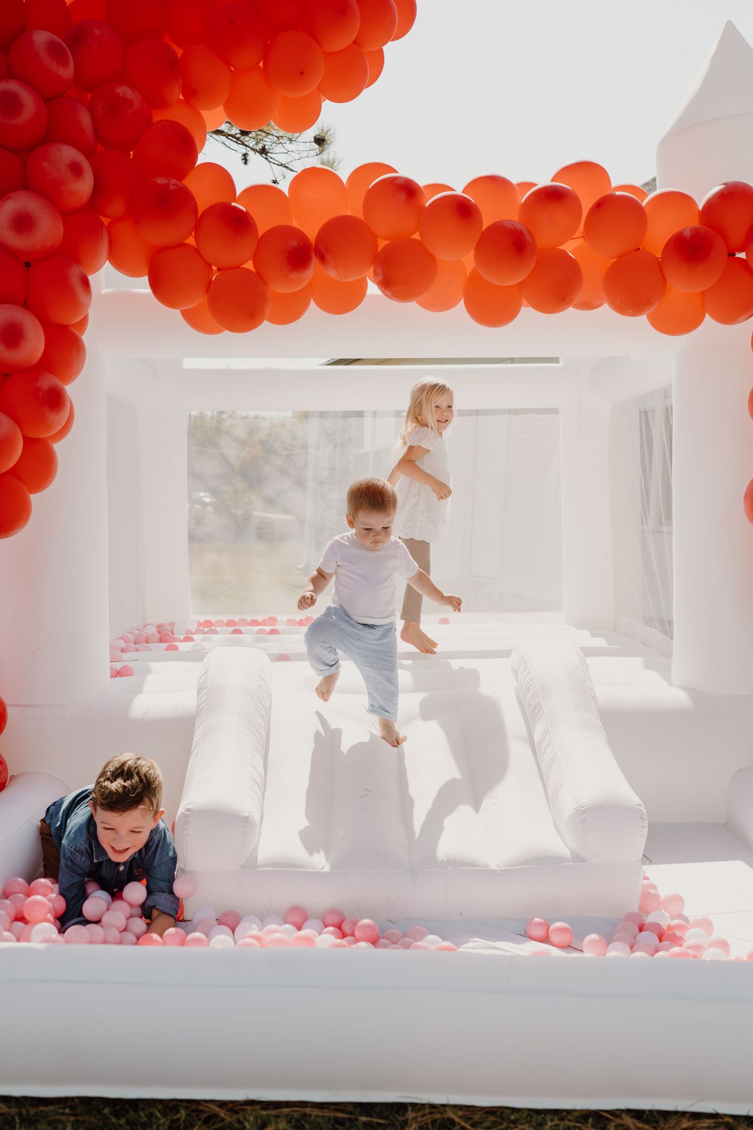 Children playing in a white bounce house decorated with red balloons and pink ball pit.