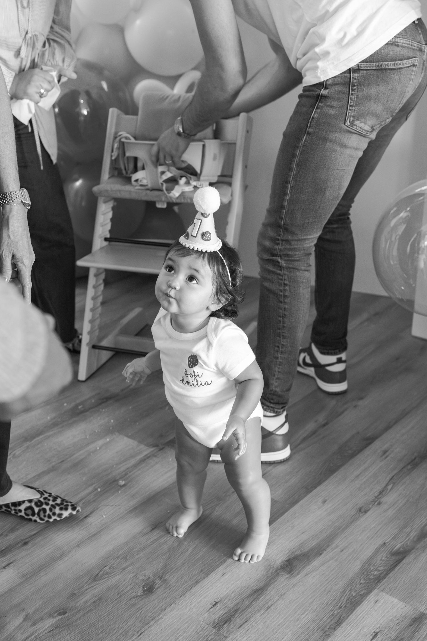 A toddler in a party setting with peach balloons and wooden flooring.