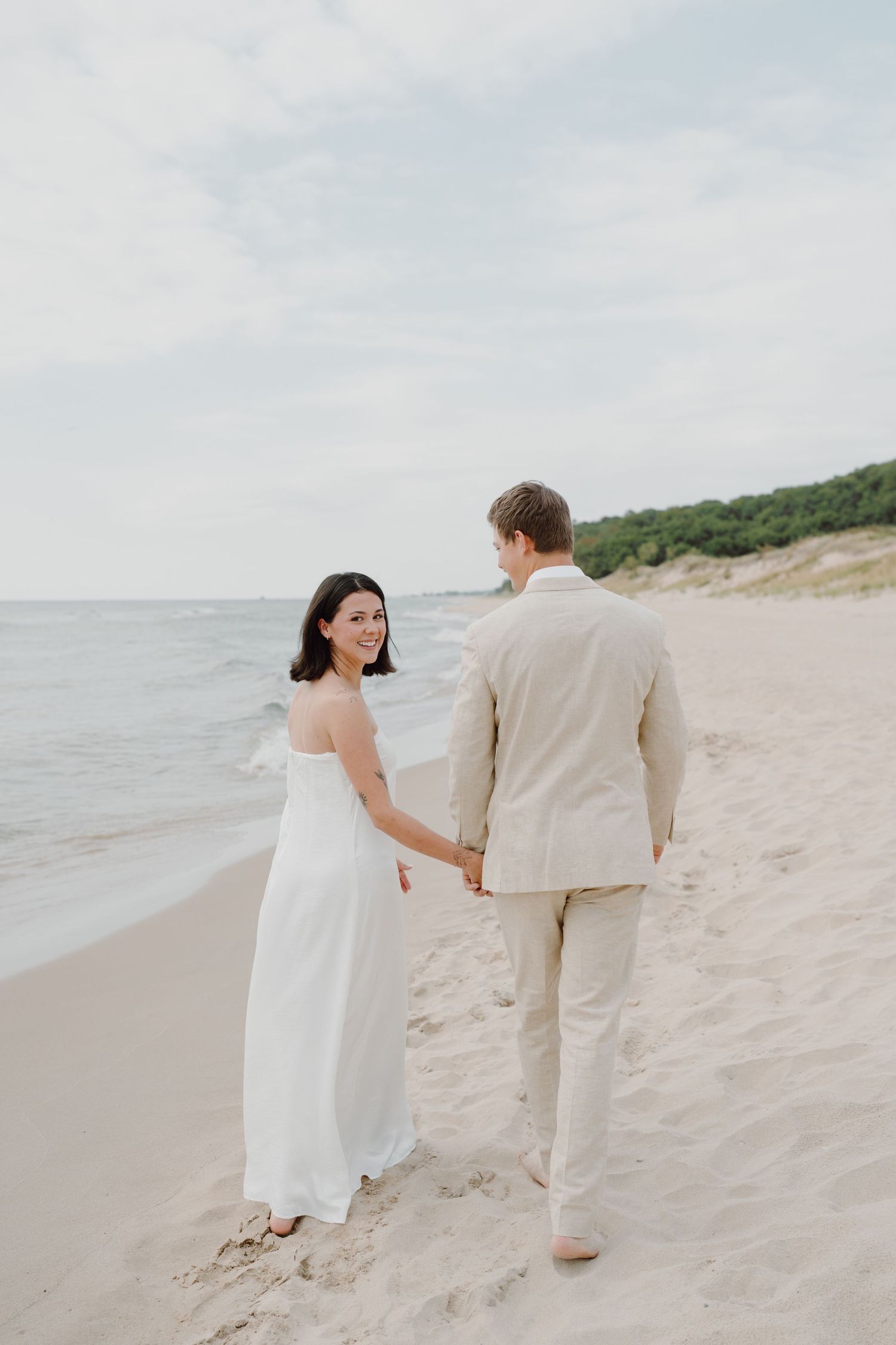 A couple walks hand in hand on a beautiful beach with sand dunes in the background.