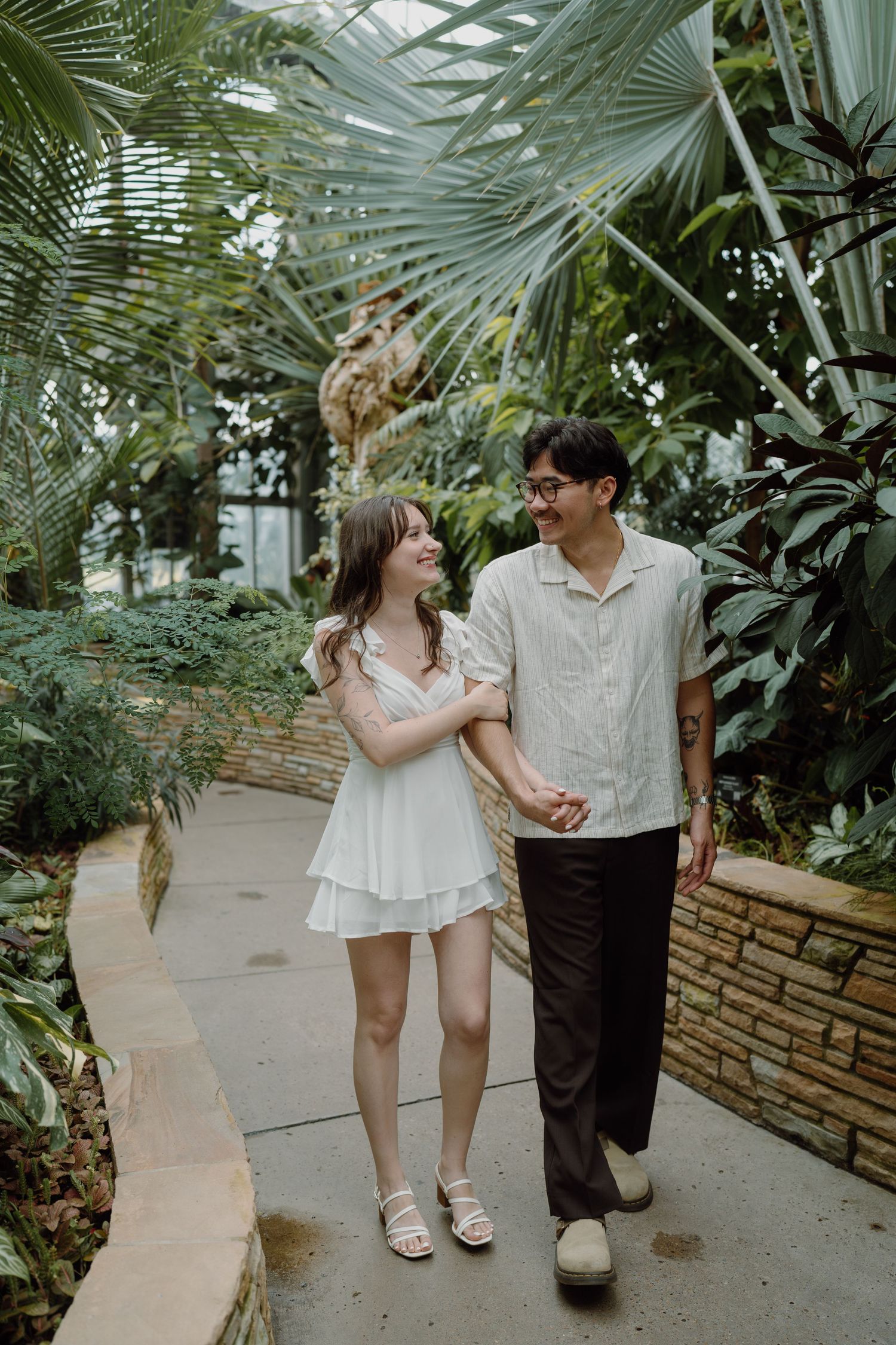 A couple walks hand in hand through a tropical greenhouse with palm trees overhead.