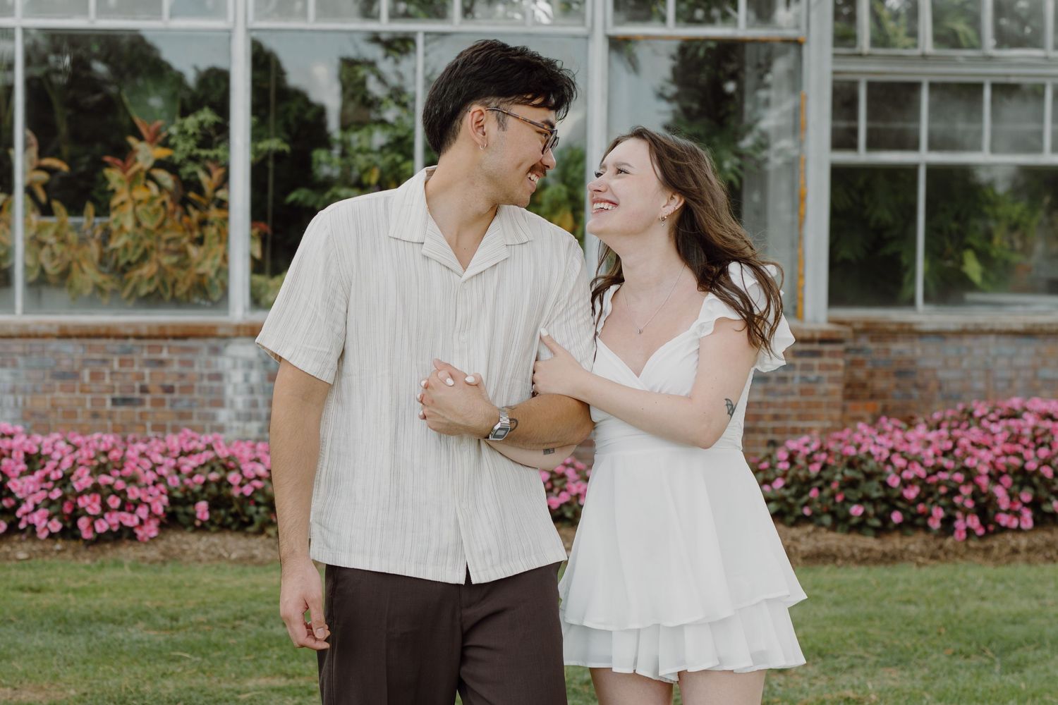 A couple shares intimate moments while posing in front of a greenhouse surrounded by pink flowers.