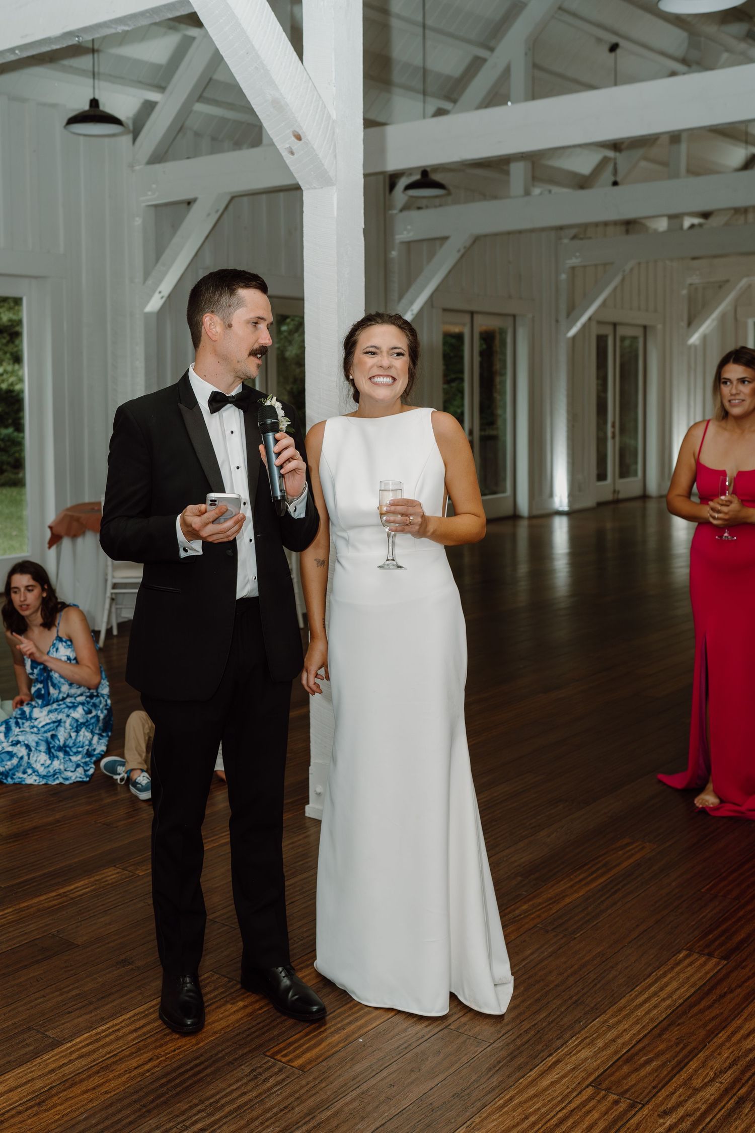 Elegant couple sharing laughs while holding champagne in a bright white barn reception venue.