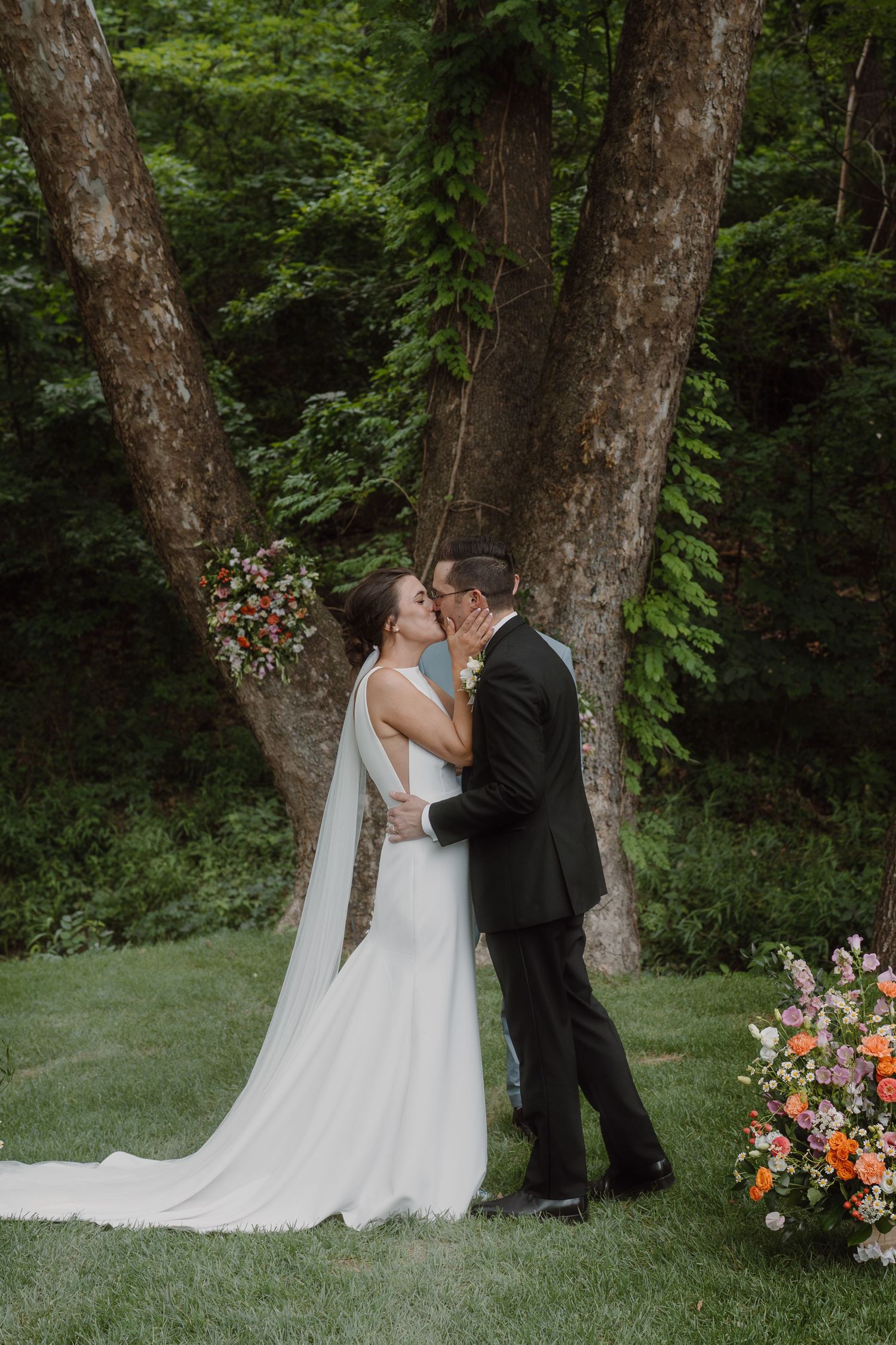 Couple sharing their first kiss as newlyweds beside vibrant floral arrangements in a forest setting.