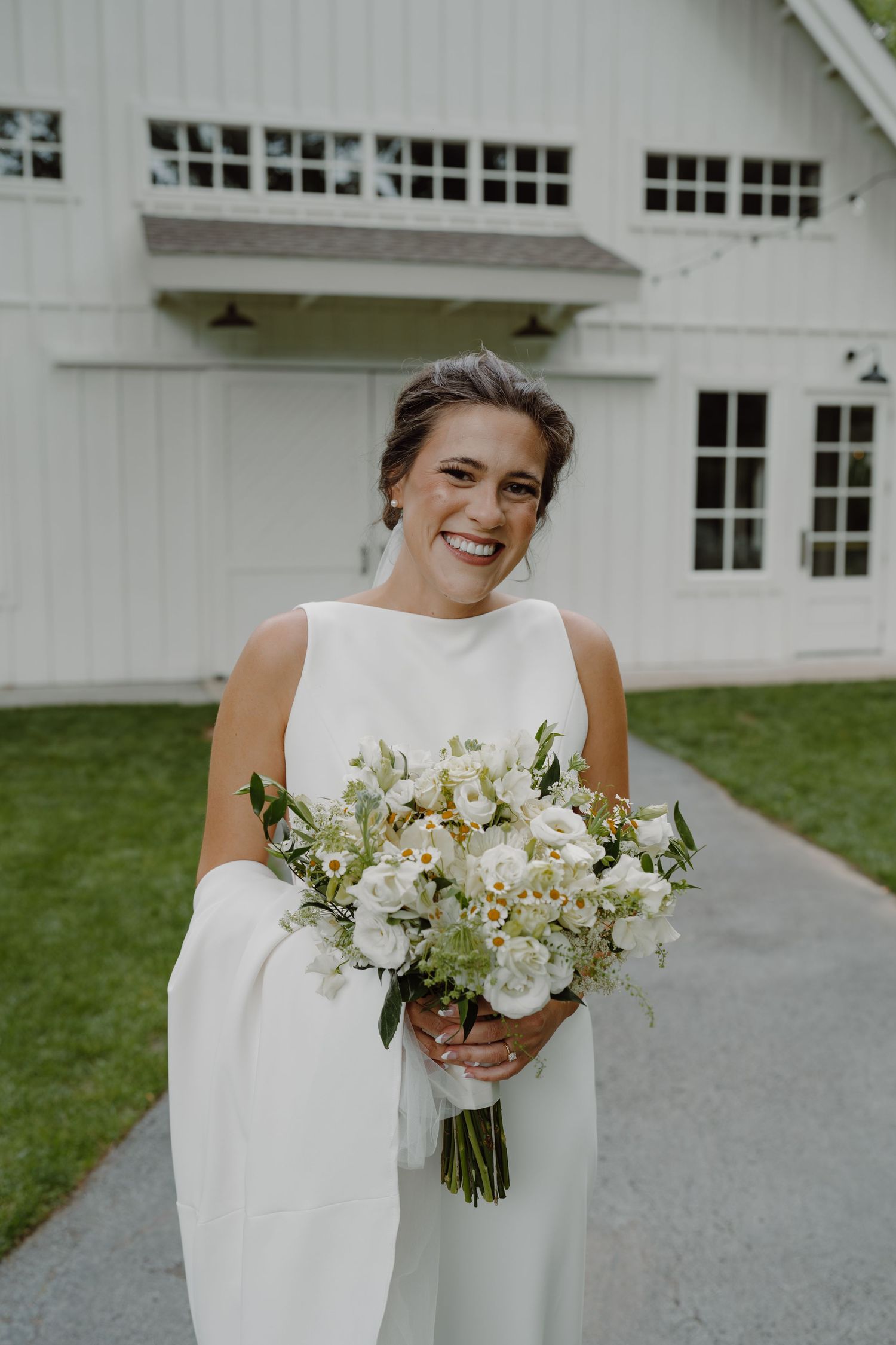 A bride holding a white and green bouquet stands in front of a white barn-style building.