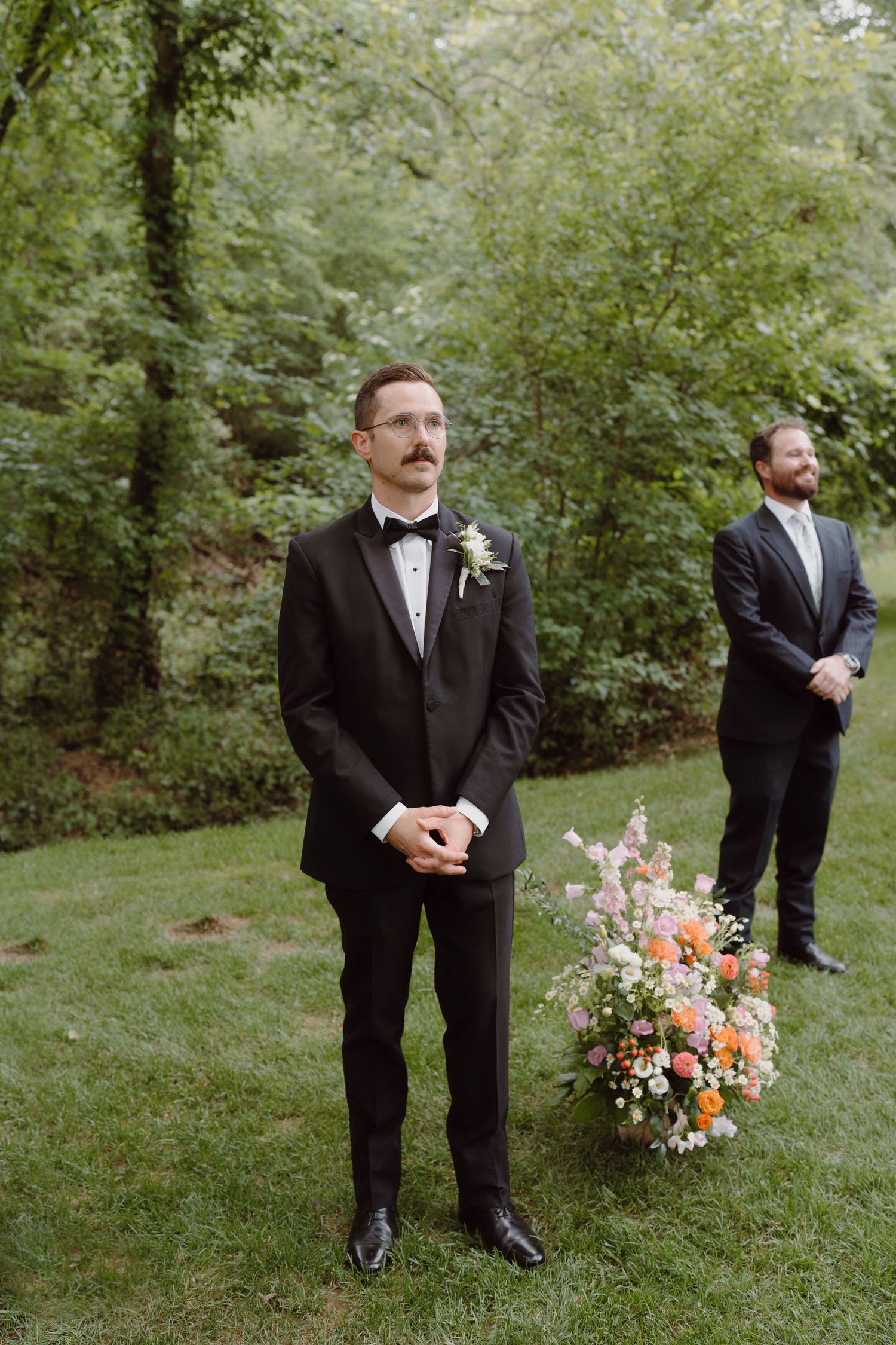 Groomsmen in black tuxedos stand spaced apart on green lawn with floral arrangements.