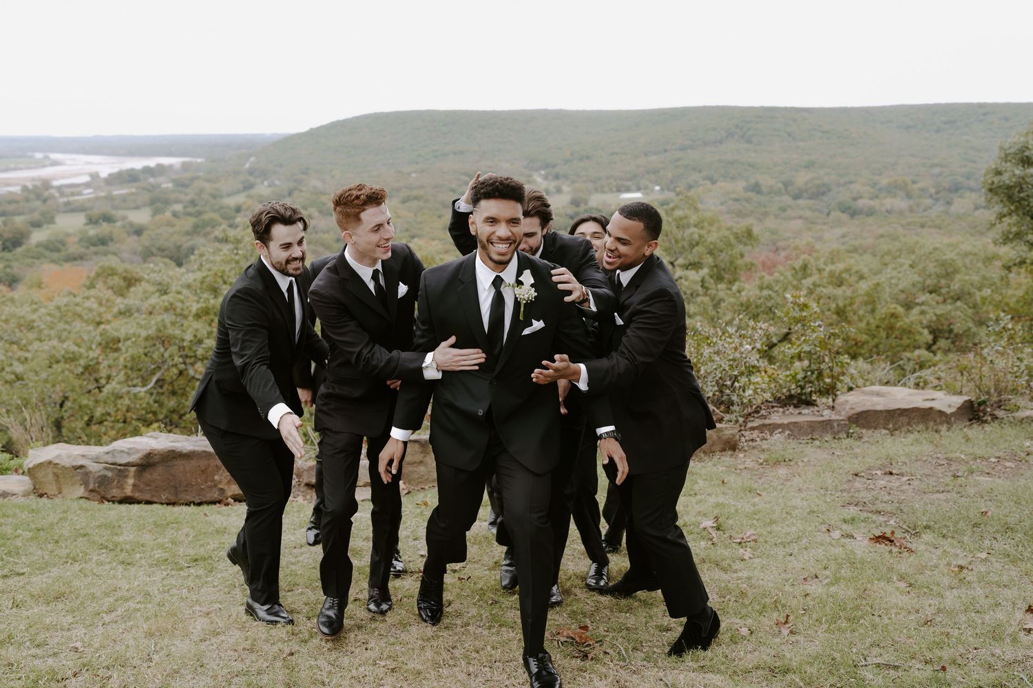 Wedding party members in formal attire leap and pose energetically against a scenic mountain backdrop.