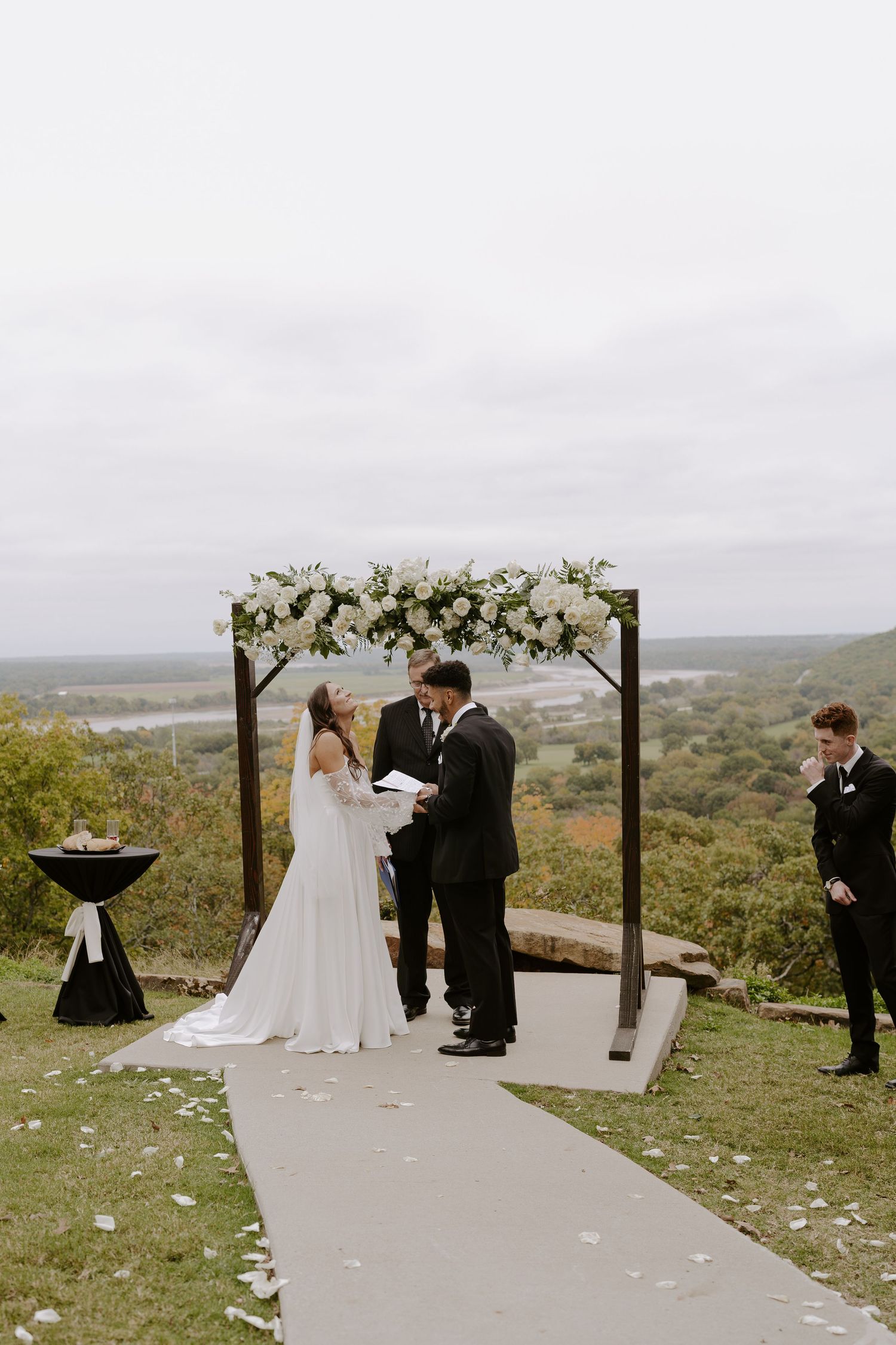 Wedding ceremony at scenic overlook with floral arch and concrete pathway against rolling hills landscape.
