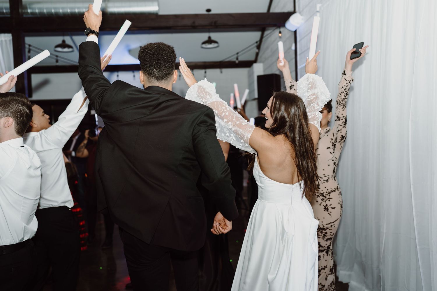 Wedding guests dance joyfully with raised arms at a reception celebration.