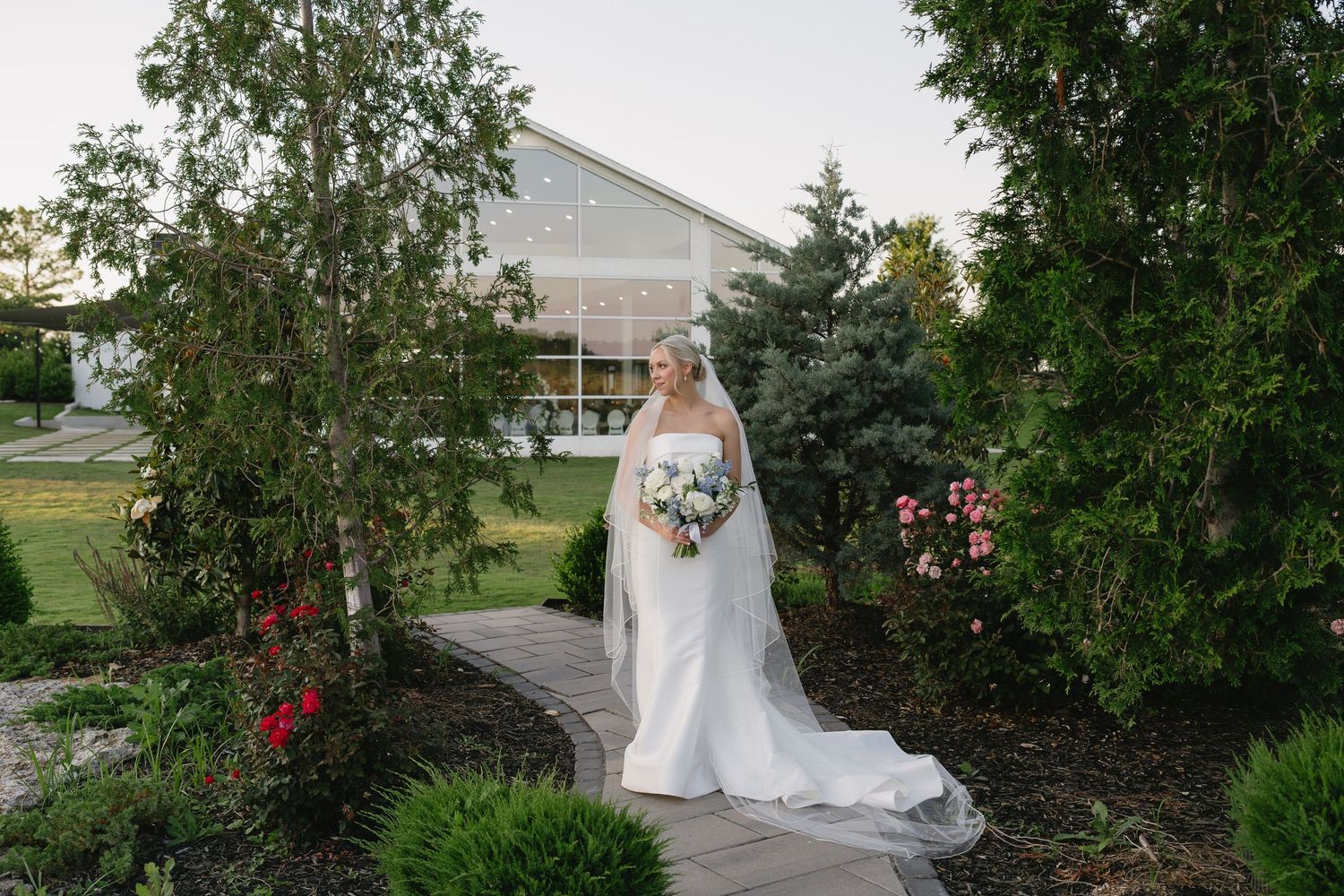Bride in white gown and veil holding bouquet on garden pathway surrounded by lush greenery near elegant venue.