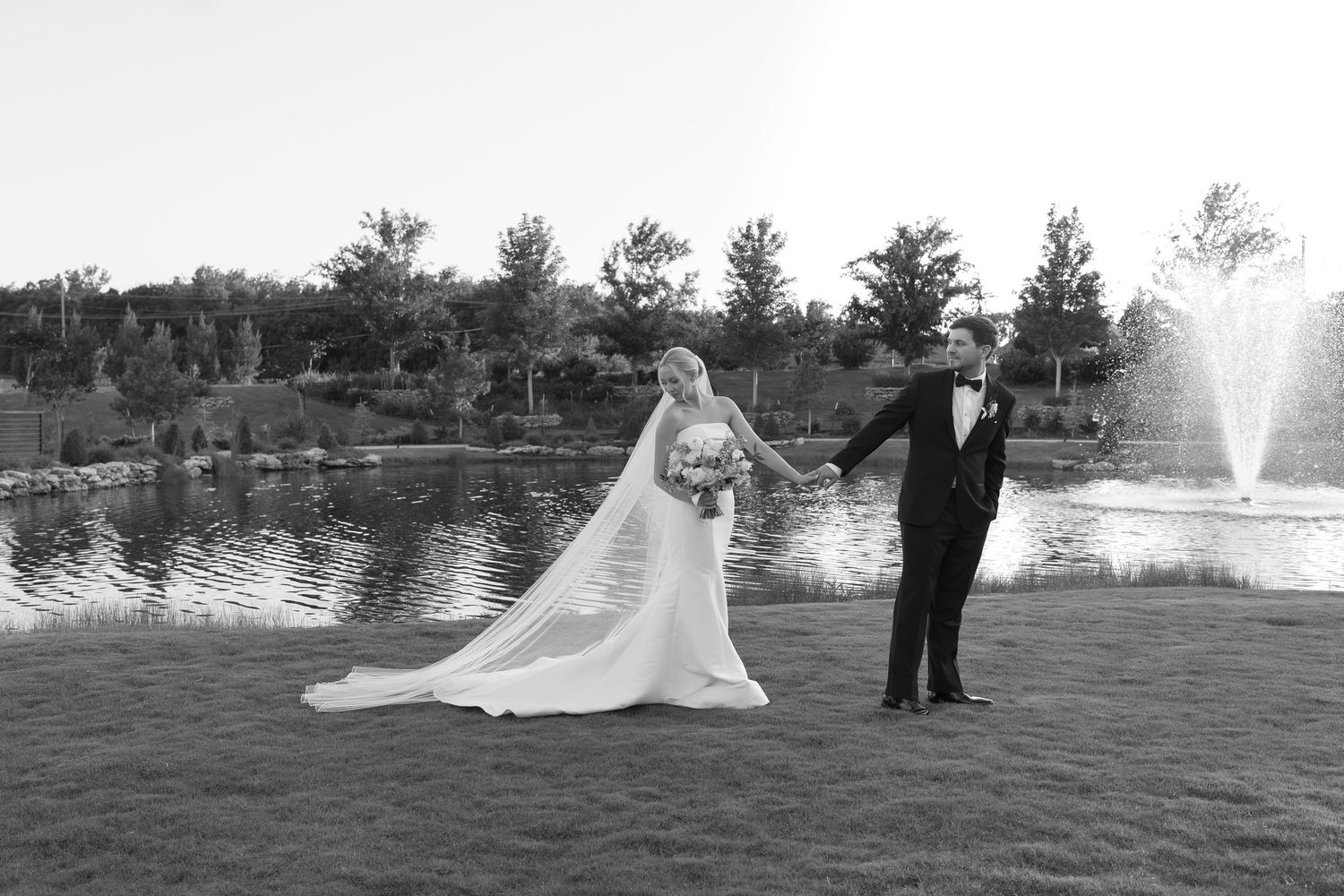 Black and white wedding photo of bride and groom holding hands by a pond with a fountain and trees in background.